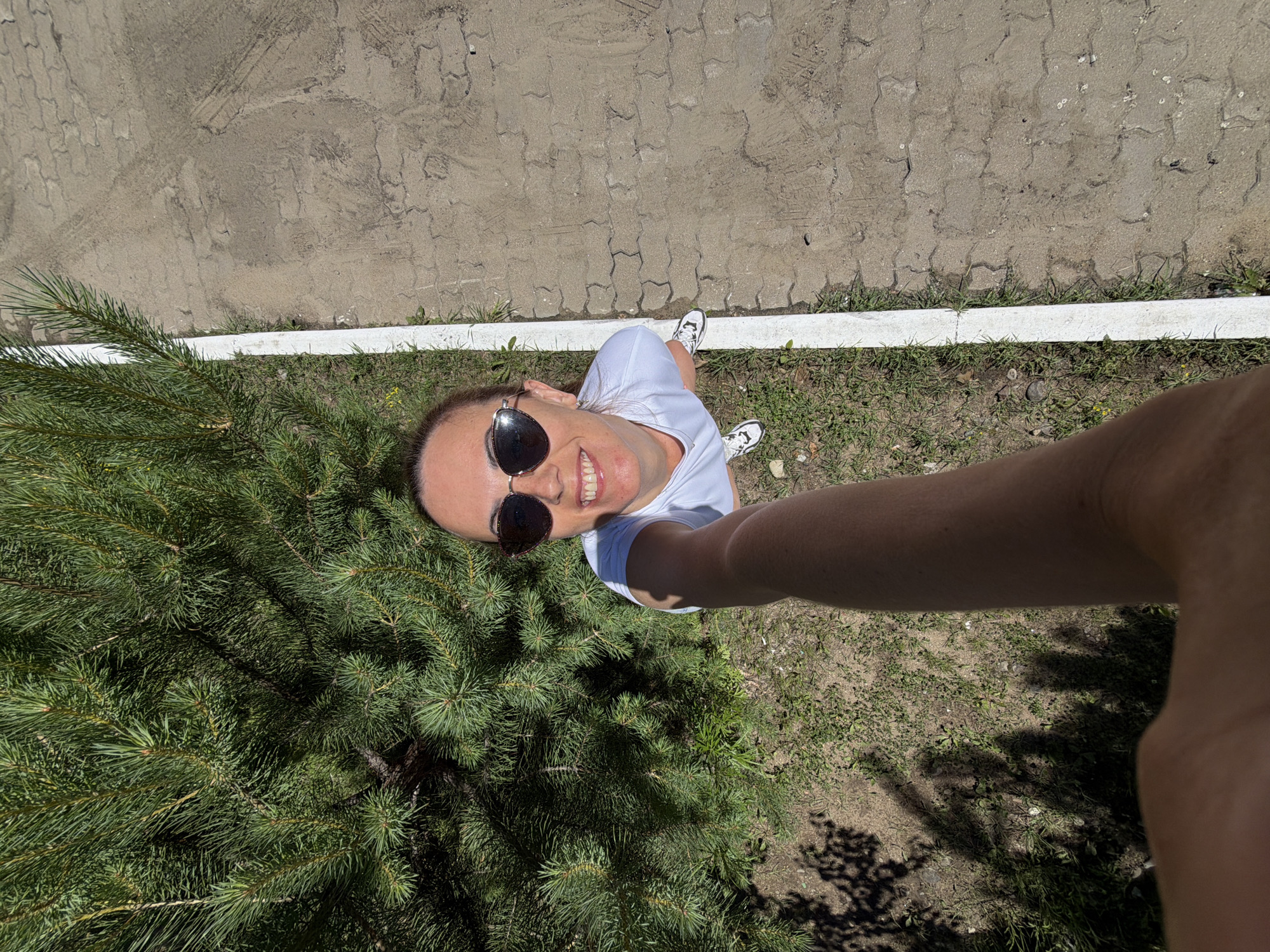 An overhead, selfie-style shot captures a Caucasian woman in sunglasses and a white t-shirt, smiling as she holds the camera. She is standing next to a lush green pine tree. To her right, a pathway of interlocking pavers is visible, bordered by a white line and grass. The ground is a mix of soil, grass, and pavers, with dappled sunlight creating shadows. The image appears to be taken outdoors in a public area, possibly a park or a paved walkway, on a sunny day.