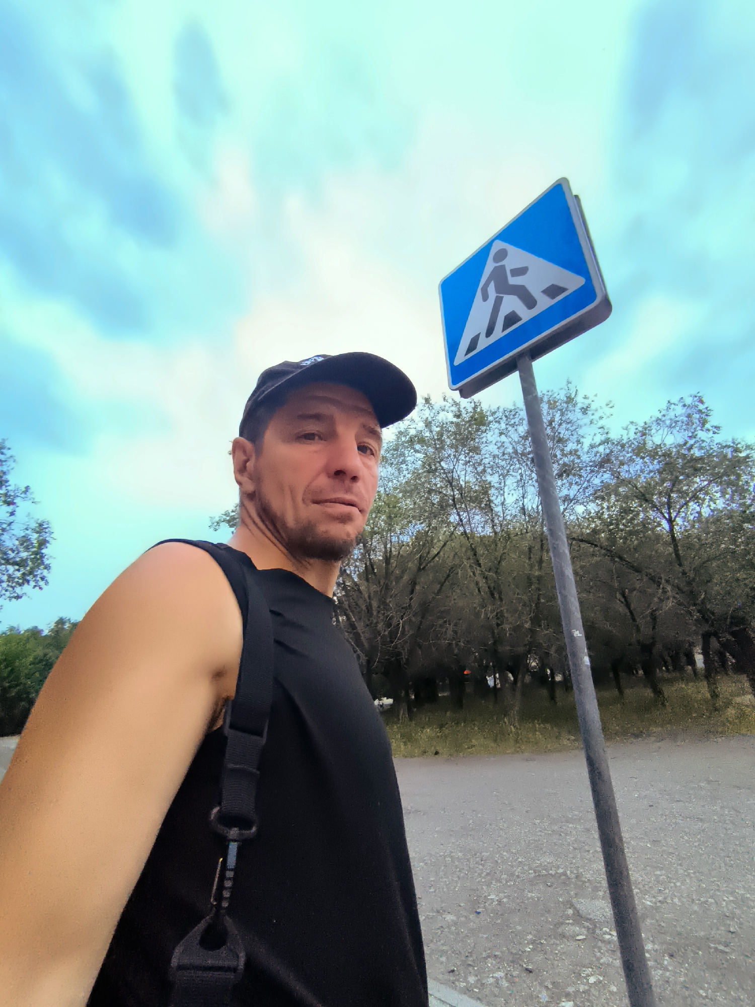 In an outdoor scene at what appears to be a pedestrian crossing in Qarağandı city administration, Kazakhstan, a man wearing a black baseball cap and a black sleeveless shirt is captured in a selfie. He is positioned in the foreground, looking towards the camera. Beside him, a blue pedestrian crossing sign with a white walking figure is prominently displayed on a metal pole. The background consists of a dirt road, green grass, and a dense line of trees under a cloudy blue sky. The lighting suggests it is daytime.