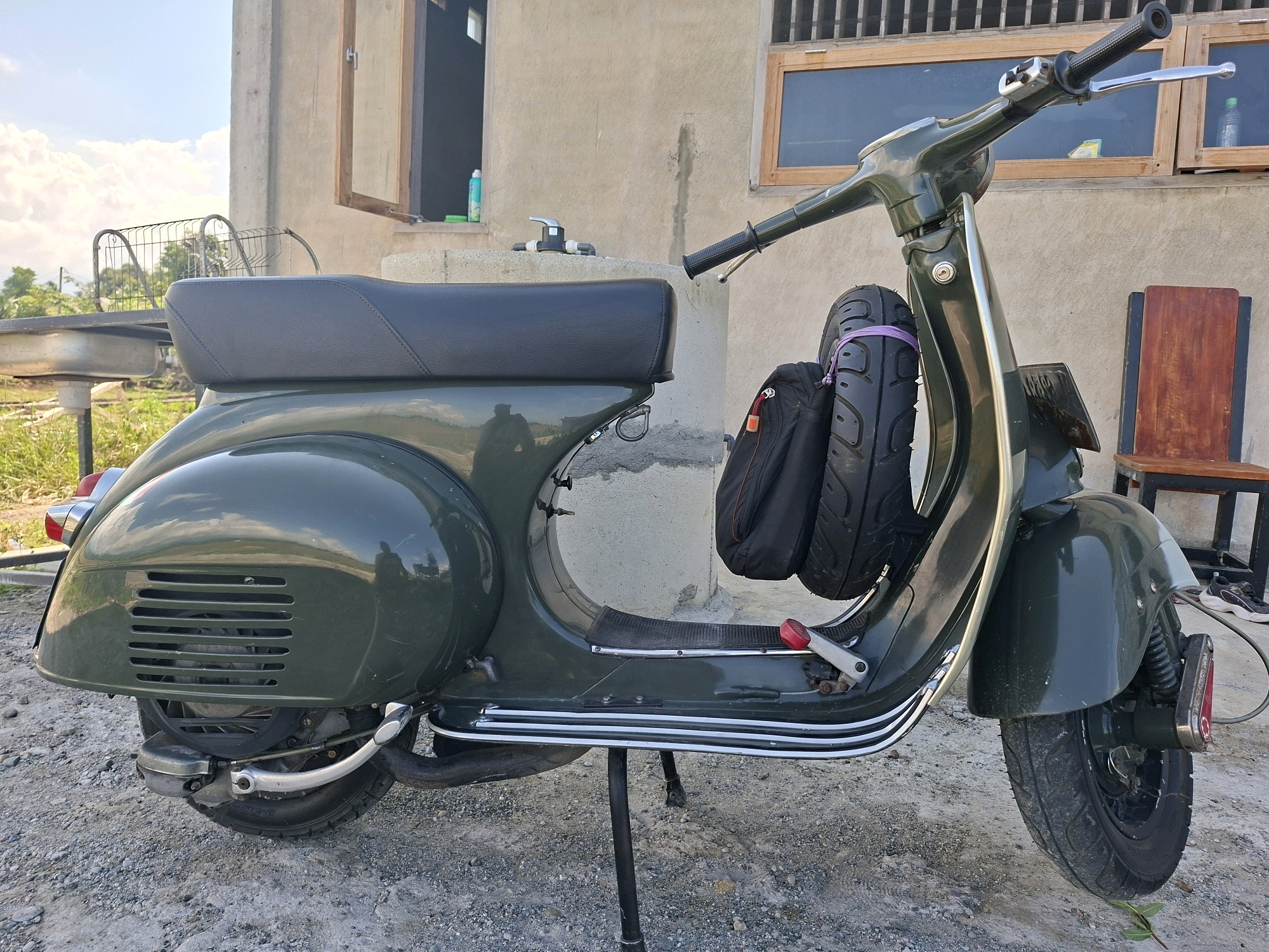 The image depicts a vintage, dark green Vespa scooter parked outdoors in Gronggrong, Indonesia. The scooter is the primary subject, positioned on a gravel surface. It is equipped with a black seat, a spare tire and a black bag strapped to the rear right side. A stand is deployed, supporting the scooter. In the background, a light-colored building with wooden window frames and a partially open door is visible. To the left of the scooter, a metal rack and a sink are present, suggesting an outdoor kitchen or utility area. A wooden chair with a red seat is situated to the right of the scooter, behind it. The sky is blue with scattered clouds. The scene appears to be in a residential or semi-rural area. No people are visible in the image.