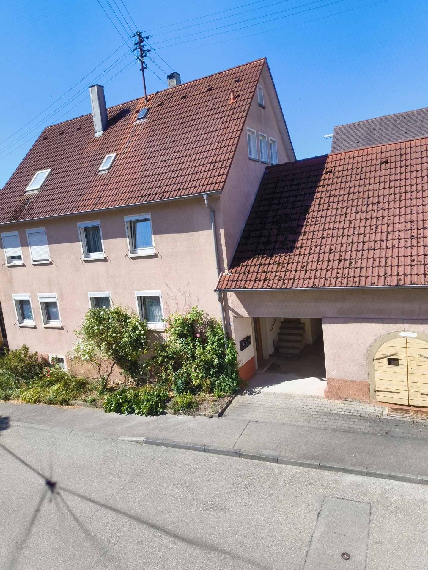 The image shows a residential street in Hardthausen am Kocher, Germany, on a clear, sunny day. A two-story building with a red tiled roof dominates the center and right side of the frame. The building has a light pink facade with multiple windows, some featuring white rolling shutters. A smaller, darker-roofed building is visible behind the main structure, partially obscured.
In front of the main building, a driveway leads to an arched opening, revealing a set of concrete stairs ascending into the building. To the right of the opening, there is a large, light-colored double wooden door with black hardware. Lush green foliage and flowering bushes are planted along the base of the building, extending towards the street.
The street in the foreground is paved with asphalt and has a raised concrete curb. The shadow of a utility pole and wires stretches across the road, indicating the presence of electrical infrastructure. The sky is a bright, clear blue. No people or vehicles are visible in the image.