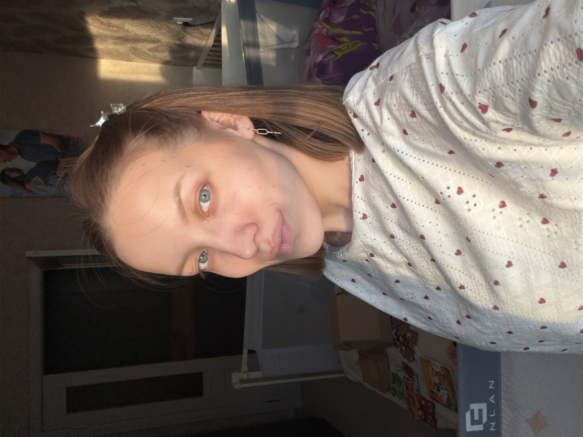 A woman with light brown hair and blue eyes is pictured indoors. She is wearing a white top with red hearts and a gold rectangular earring. The background suggests a bedroom or living space with a window, a patterned blanket, and furniture. A hair clip is visible in her hair. There is no information to suggest that the image was taken in Reshetnikovo, Russia, or any other particular location.