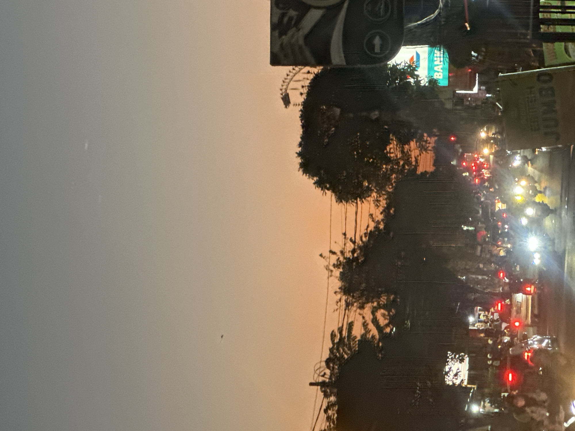 The image depicts a street scene in Denpasar, Indonesia, at dusk. A traffic jam is evident with numerous motorcycles and a few cars on the road. The sky exhibits a gradient of orange and grey, suggesting the setting sun. Silhouettes of trees line the street, and power lines are visible. A large billboard with "BANK" written in green letters is prominent on the right side of the frame. Another sign with "JUMBO" in yellow is also visible. Some individuals are visible on motorcycles, many wearing helmets. The overall atmosphere is that of a busy urban environment transitioning into nighttime.