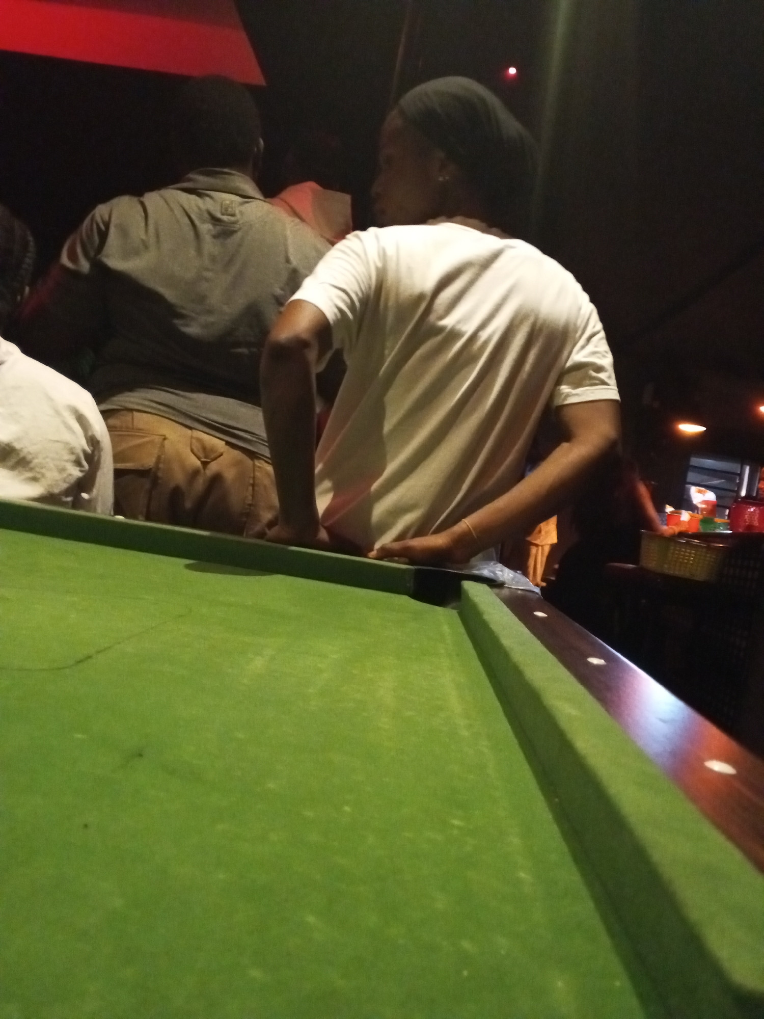 In a dimly lit indoor setting in Lokoja, Nigeria, the focus is on a green-felted pool table. Two individuals are positioned on one side of the table. One person, wearing a gray shirt and khaki pants, stands with their back to the camera. The other, closer to the viewer, is dressed in a white t-shirt and has a black head covering. Their hand rests on the edge of the pool table. Behind them, other figures are visible, along with various objects including containers and what appears to be a window. The environment suggests a bar or similar entertainment venue.