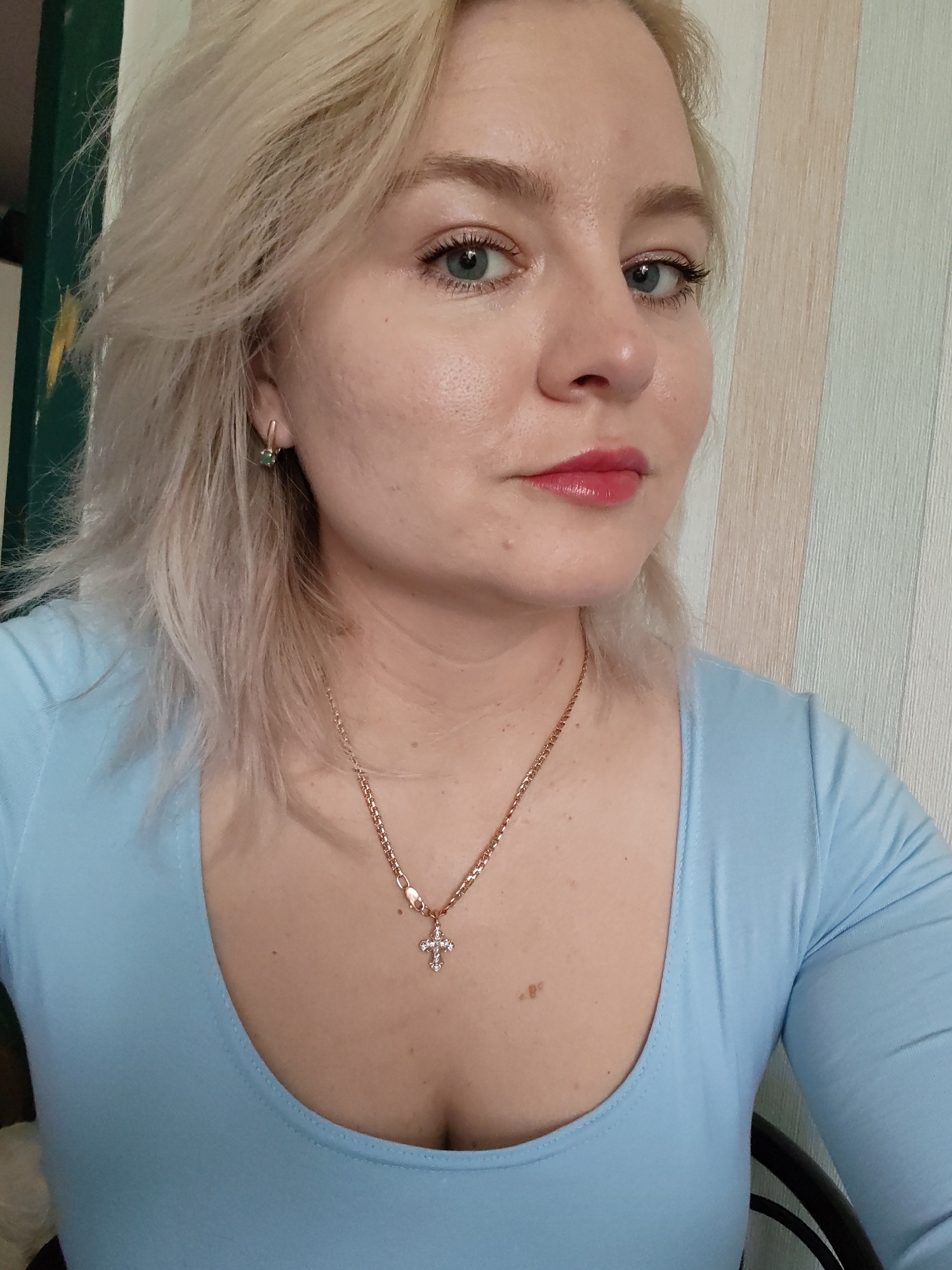 A blonde female, estimated to be in her late 20s to early 40s, is shown in a chest-up portrait in Izhevsk, Russia. She has fair skin, light blue eyes, and shoulder-length blonde hair. Her lips are coated with a glossy, reddish-pink product. She is wearing a light blue, long-sleeved top with a scoop neckline. A small, rose gold colored hoop earring with a dark green gemstone is visible in her right ear. Around her neck, she wears a rose gold colored chain necklace with a small, clear-stoned cross pendant. The background on the left side of the frame features a dark green vertical surface adjacent to a thin, lighter vertical strip. On the right, the background consists of vertically striped wallpaper in light tones of off-white, beige, and soft green.