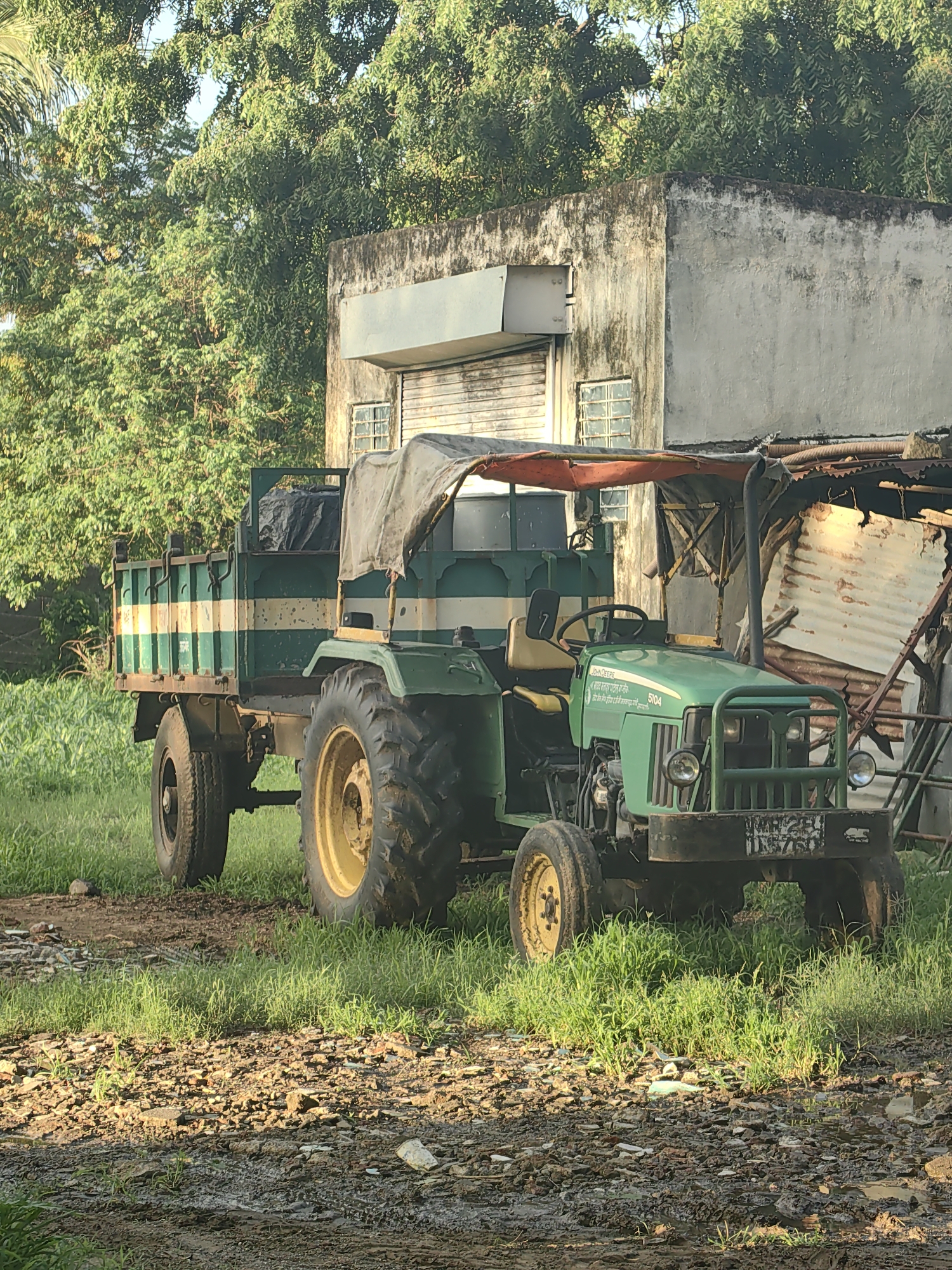 A green tractor with a trailer is parked on a dirt and grass area in front of an old concrete building with a corrugated metal roof. The trailer is green and white striped and loaded with black material. The tractor has a yellow canopy and a black exhaust pipe. There is a license plate on the front of the tractor that reads "MH29 F 7576". Trees are visible in the background. The scene appears to be in Mhaiswadi, India.