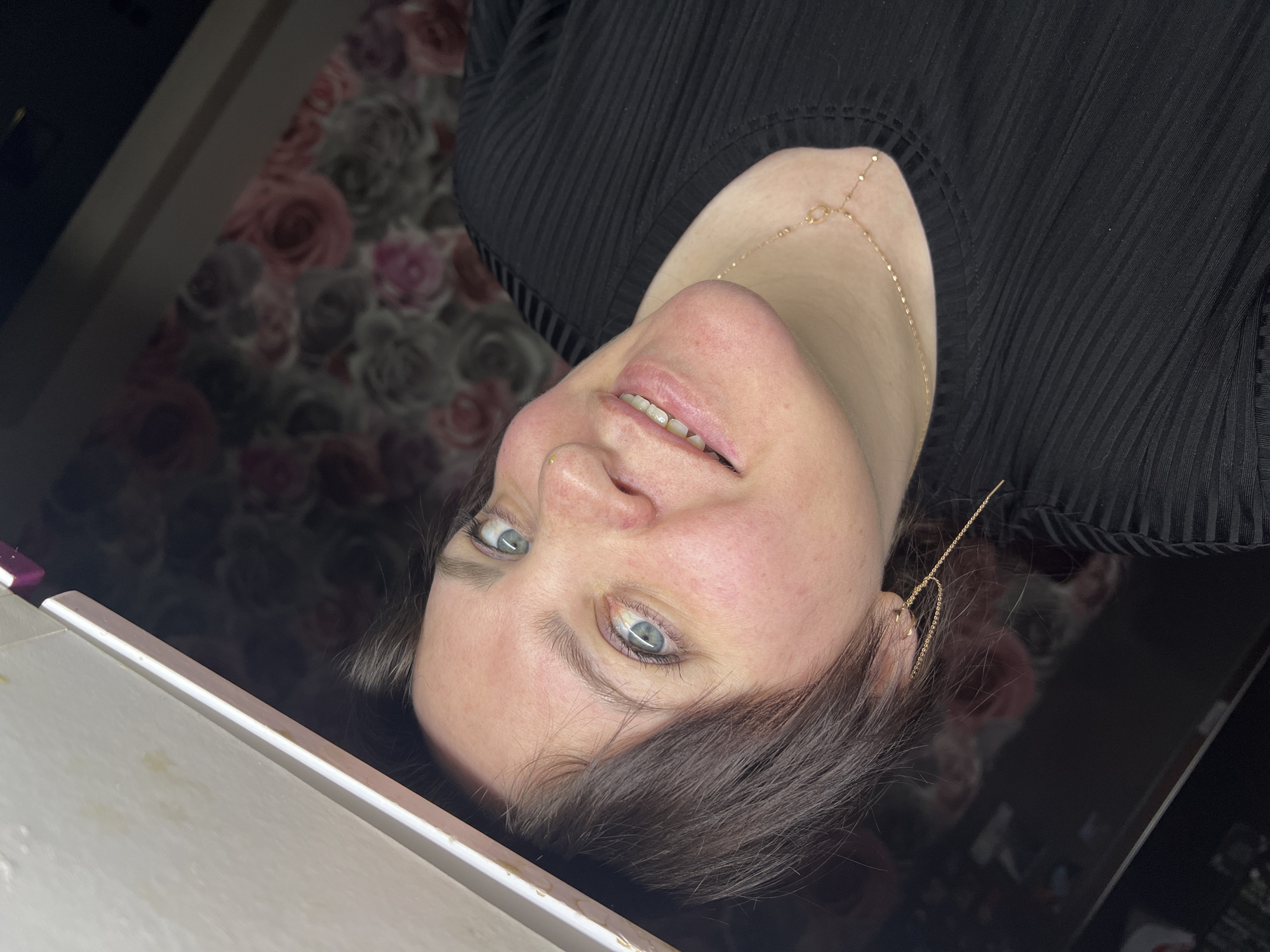 A woman is lying on her back. She has fair skin, light blue eyes, and short, dark brown hair. She is wearing a black, ribbed top and a delicate gold necklace and earrings. The background includes a floral patterned wall and a white surface with a glossy dark surface adjacent to it. There is no information about a location in Noviy, Russia in the image.