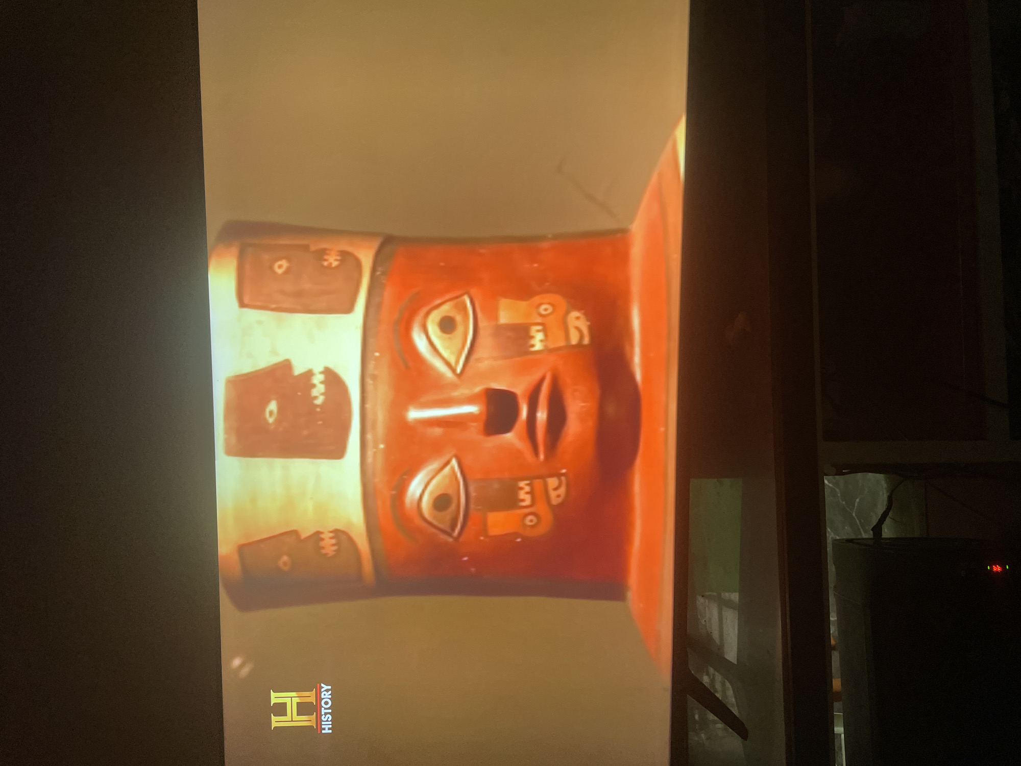 The image shows a red artifact with a face on it, likely a vase or sculpture. The face has stylized features and is decorated with patterns or designs. There are additional rectangular sections with designs on the upper part of the artifact. The artifact is displayed on a screen displaying a History channel logo. It is filmed inside a room at night.