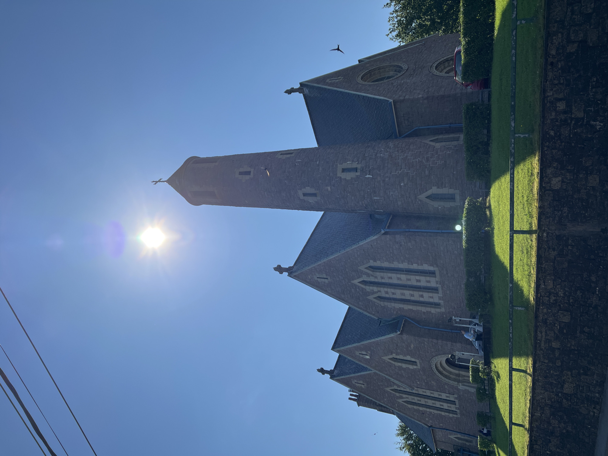A stone church with a tall, cylindrical tower topped with a cross stands under a bright, sunny sky. The sun is visible in the upper portion of the image, creating a lens flare. The church has multiple pointed gables with dark, sloped roofs and arched windows with decorative stonework. A smaller cross is visible on the peak of one of the gables. In front of the church, a well-maintained lawn slopes down to a stone wall, with hedges planted along the base of the church. A few people are visible on the lawn near the church entrance, and a red car is parked to the right of the building. Several birds are in flight in the sky. The scene appears to be set in a suburban or semi-rural environment. The location is not specified as Donegal, Ireland.