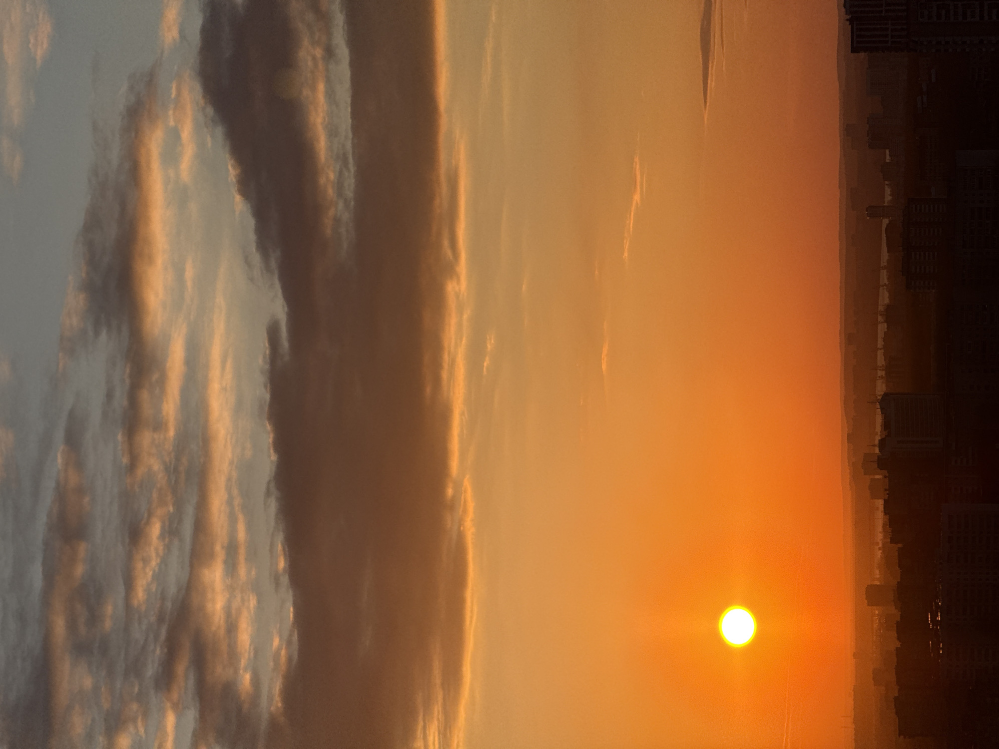 A vibrant sunset graces the sky over the city of Perm, Russia, captured from an elevated perspective. The sun, a brilliant, nearly white orb with a faint flare, is positioned low on the horizon towards the bottom right of the frame. Its intense light bathes the lower atmosphere in vivid oranges, yellows, and reds.
The sky directly surrounding the sun is a bright orange, gradually softening to an amber hue, then transitioning to a muted blue-gray towards the upper left. This upper portion of the sky is adorned with diverse cloud formations. Some appear as dark, textured masses, silhouetted against the brighter sky, while others are illuminated from below, revealing golden and orange accents.
Along the bottom and left side of the image, the city's extensive skyline forms a dark silhouette against the radiant backdrop. Numerous buildings and structures are discernible, creating a dense urban tapestry that extends into the hazy distance. There are no identifiable people, specific landmarks, or visible text. The overall scene conveys the serene and awe-inspiring beauty of dusk over a sprawling metropolis.
