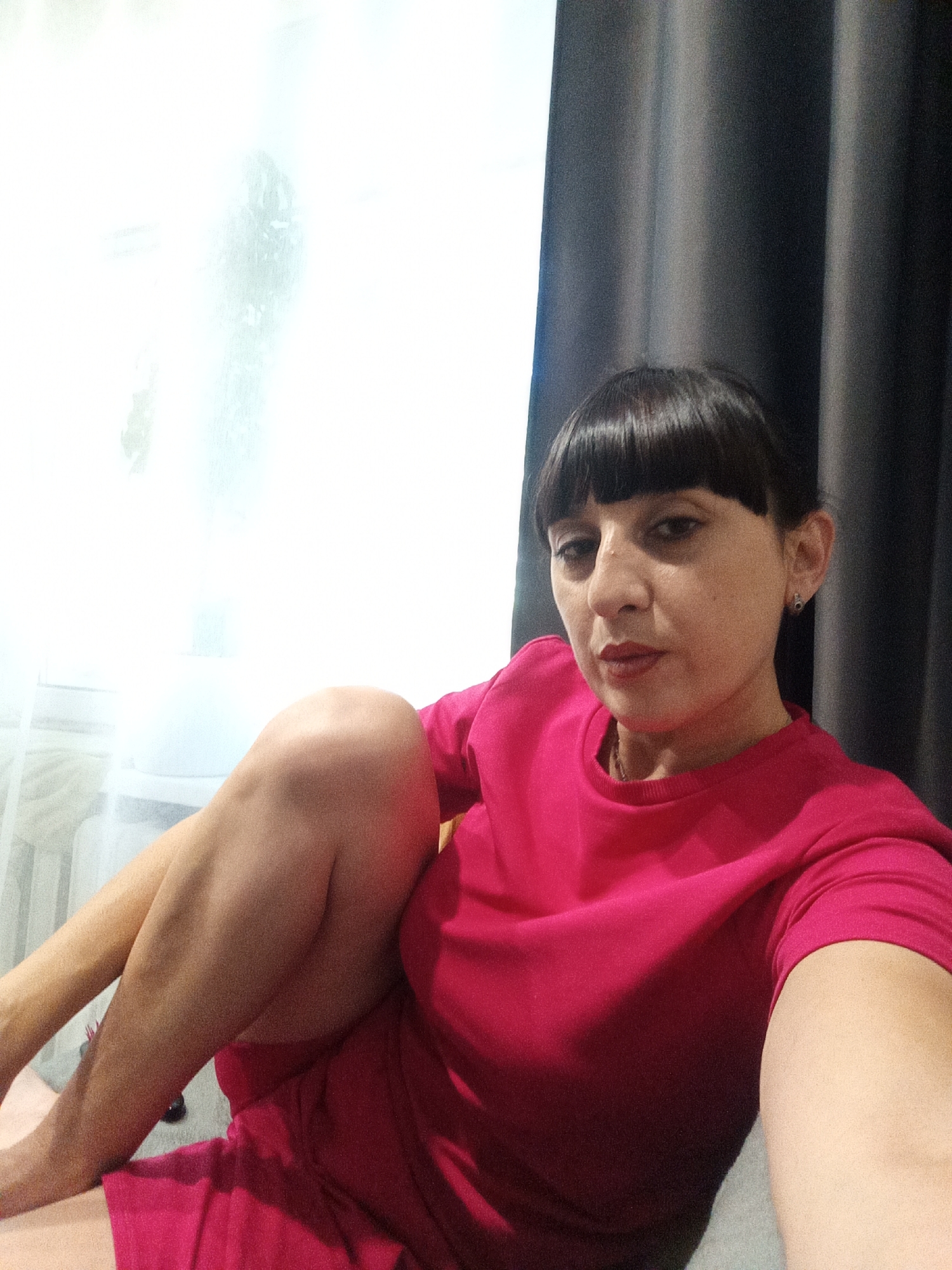 A medium shot captures a woman with dark hair and bangs, wearing a bright pink sweatshirt. She is seated, with her legs crossed, and her right arm is extended, holding a device that appears to be a smartphone, suggesting she is taking a selfie. The background is divided between a bright, diffused light source on the left, likely a window with sheer curtains, and dark, vertical curtains on the right. A small, dark earring is visible in her left ear. The scene is indoors, possibly in a residential setting in Belogorsk, Russia, indicated by the context provided.