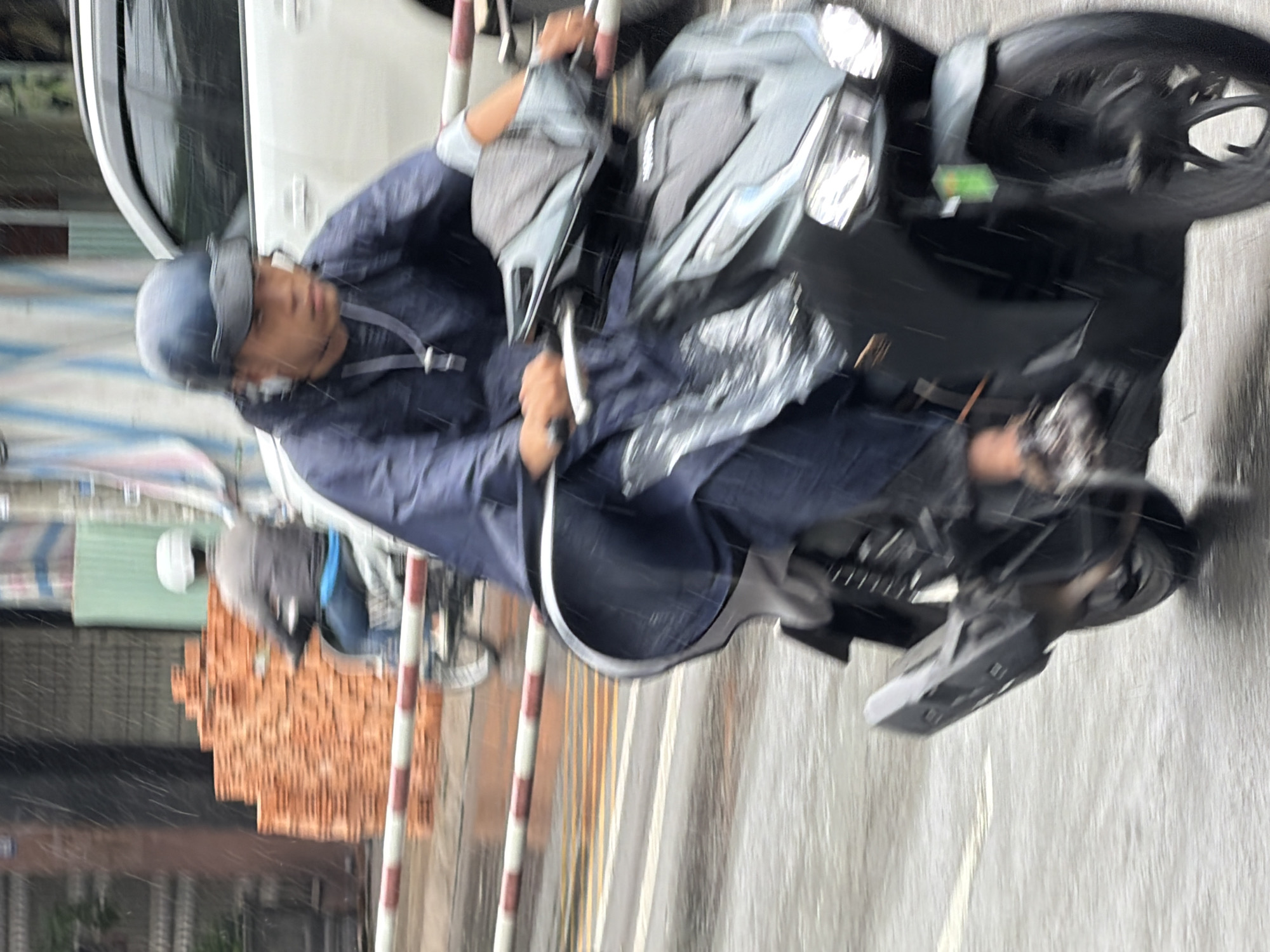 The image depicts a street scene in Nha Trang, Vietnam, during rainfall. A person wearing a dark blue poncho and a dark helmet is riding a dark-colored scooter. The person's face is visible, with a focused expression. The scooter has visible headlights and wheels, and the rider's hands are on the handlebars. In the background, a white car is partially visible, as is a stack of orange bricks and a pedestrian wearing a white helmet on a bicycle. The street is wet and reflective, indicating the rain. Red and white striped barriers are present in the foreground and middle ground, suggesting a road closure or construction area. The overall impression is a dynamic, motion-blurred snapshot of urban life during inclement weather.