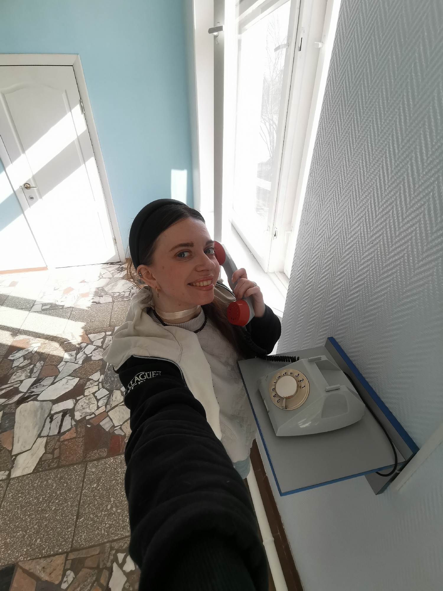 A young woman with dark hair pulled back by a black headband is the central subject of this high-angle selfie. She is smiling broadly at the camera, holding a vintage rotary phone receiver with a red earpiece to her left ear. Her right arm extends towards the camera, suggesting she is taking the photo herself. She wears a light-colored top under a black and white hooded jacket, with visible text "LEAGUE OF" on her left sleeve. A thin, light-colored choker is around her neck.
She is standing indoors, in what appears to be a hallway or corridor. The walls are painted a light blue, and to her right, there is a section of wall covered in light gray wallpaper with a distinct herringbone pattern. A bright window is visible on the right, letting in strong sunlight that casts clear shadows on the light-colored mosaic-tiled floor. The floor consists of irregular pieces of various light and dark brown, beige, and white stones or tiles. A white door with a simple handle is visible to her left, also illuminated by sunlight.
The gray rotary telephone, with a cream-colored dial, rests on a light gray shelf or niche mounted on the patterned wall. Its coiled black cord is visible. The bright lighting suggests it is daytime, likely sunny. The overall atmosphere is light and cheerful, reflecting the woman's expression.