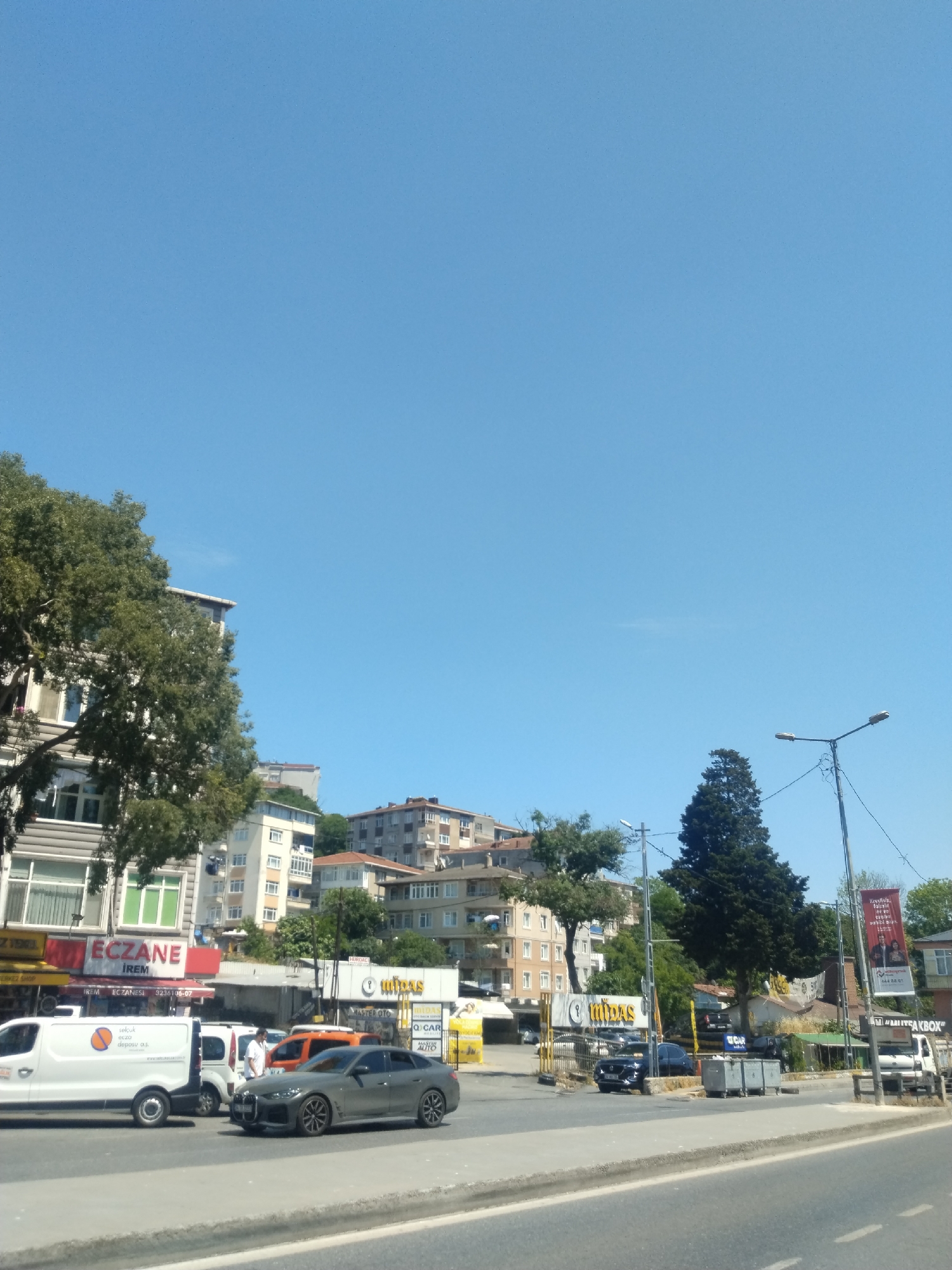 This image captures a vibrant, sunny daytime street scene in Sarıyer, Turkey. A clear, expansive blue sky dominates the upper portion of the frame.
In the foreground, a road with multiple lanes is visible, populated by several vehicles. A white delivery van with the text "selcuk ecza deposu as." and a distinctive orange and white logo is prominent on the left. Beside it, a person in a white shirt stands near a grey sedan, which appears to be a BMW 4 Series Gran Coupe. An orange car is partially visible behind the grey sedan.
To the left of the street, multi-story urban buildings are visible, interspersed with large, leafy green trees. One building features a bright red and white sign for "ECZANE İREM" (Irem Pharmacy), along with the phone number "3236106-07." Another sign above it reads "ERKEZ SHOP" (likely Merkez Shop).
On the right side of the road, another large, dark green conifer tree stands out. Further down, signs for businesses such as "MİDAS," "MASTER OTO," and "Q CAR AUTO" indicate automotive services or dealerships. Several cars, including a dark blue one, are parked in this area. Two large grey dumpsters are positioned near the roadside. A red banner displaying Turkish text, including "Kiralık" (for rent), is partially visible on the far right. Streetlights illuminate the area. In the background, more buildings are nestled on a sloping hillside, characteristic of the region. The overall impression is one of a busy urban environment on a clear, bright day.
