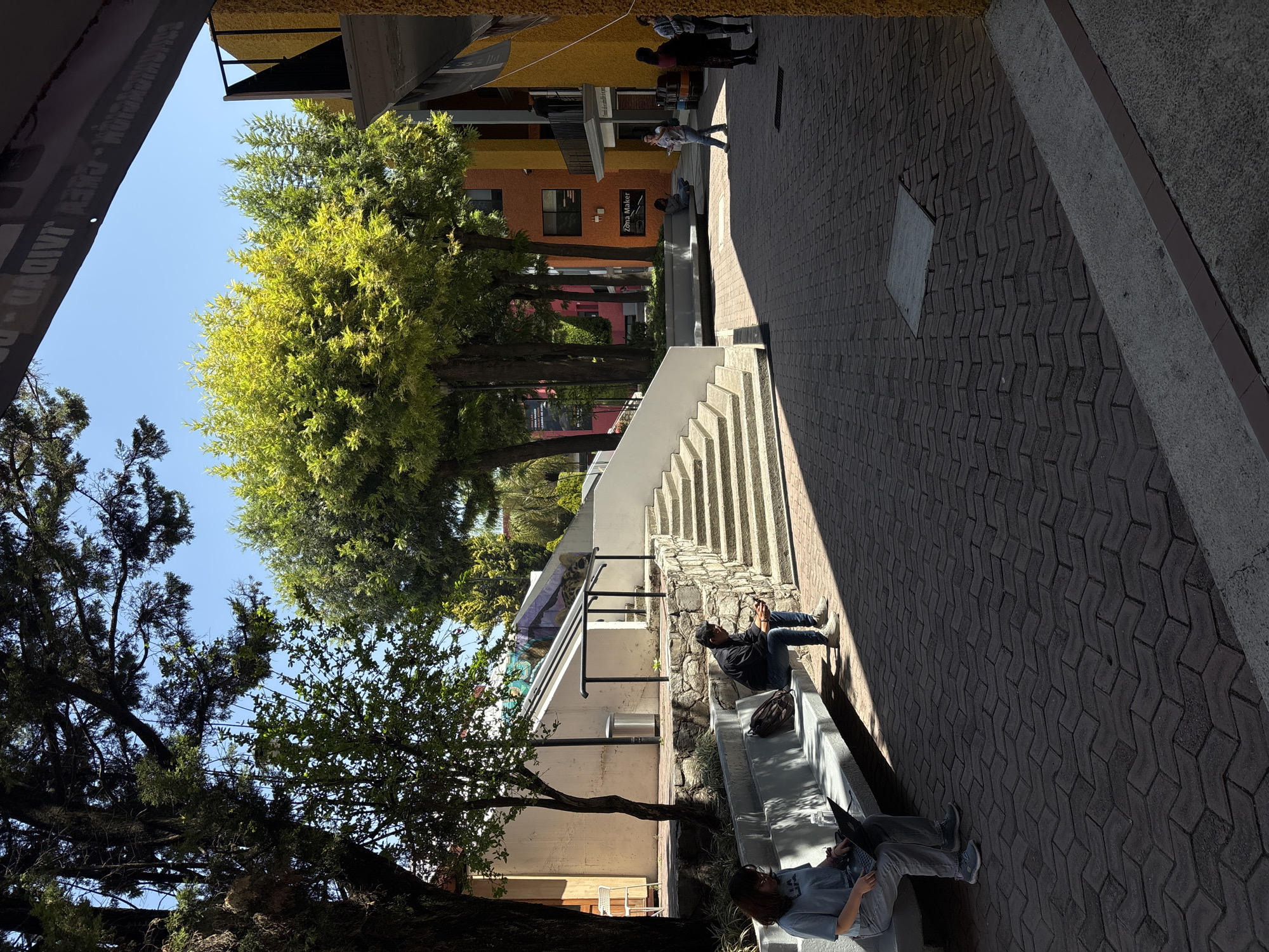 The image depicts a pedestrian walkway in San Andrés Cholula, Mexico. Two individuals are seated on a low concrete bench, one with a laptop. A person is seated on a raised ledge alongside a flight of stairs. Several people are walking in the background. The walkway is paved with interlocking bricks. Buildings of various colors line the walkway, with trees interspersed throughout. The sky is clear and blue.