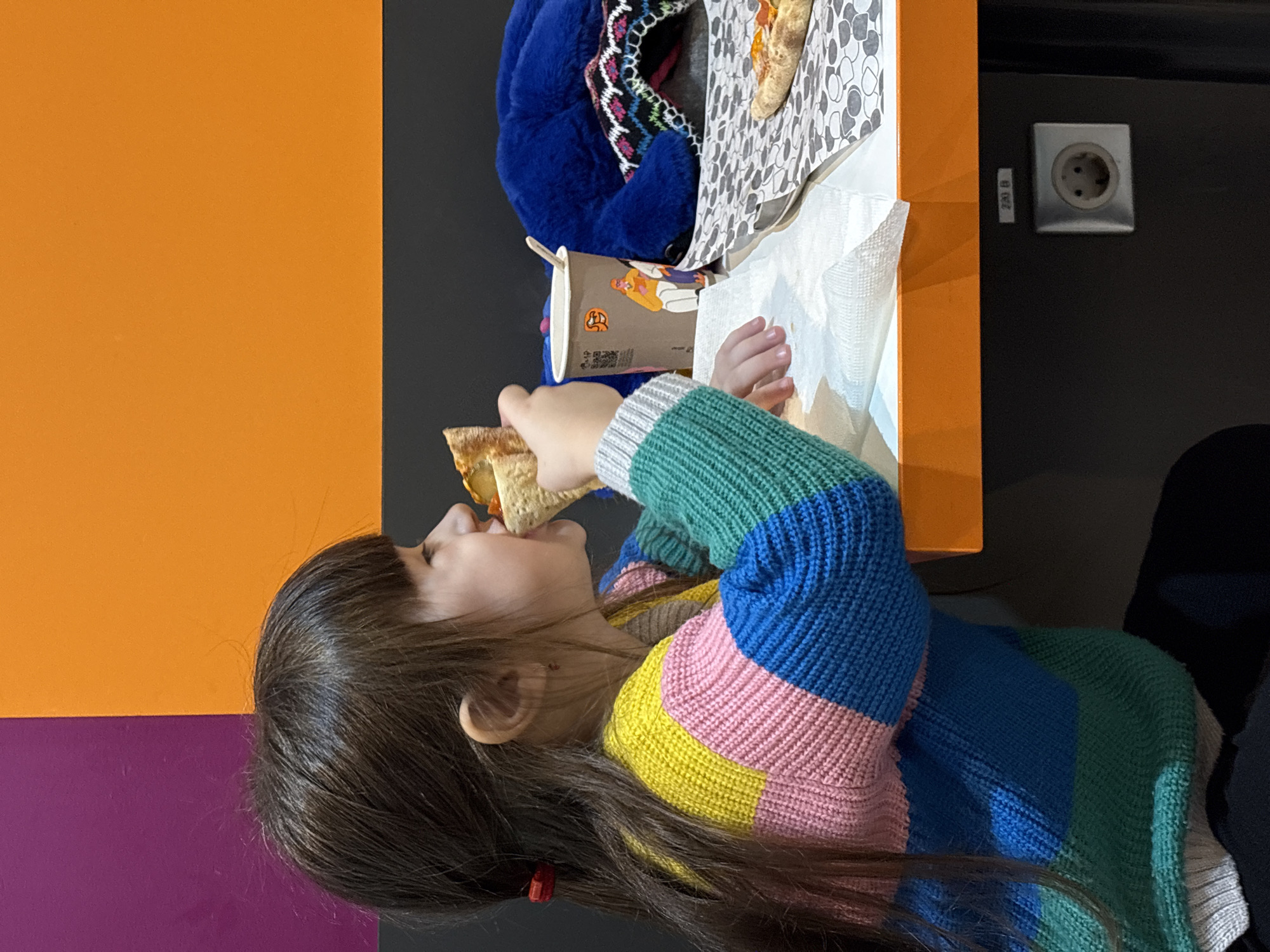 A young girl, appearing to be of elementary school age, is depicted in profile, seated at an orange-topped table, actively eating a slice of pizza. She has long, dark brown hair with bangs and wears a multi-colored knitted sweater with horizontal stripes of yellow, pink, blue, green, and light gray. A red hair tie is visible in her hair. Her right hand holds the pizza slice to her mouth.
On the orange table to her right, a brown Dodo Pizza branded paper cup with a lid and a wooden stirrer is present. White paper napkins and a piece of patterned paper liner are also on the table, supporting another partially visible pizza slice. A dark blue, possibly faux-fur, item is situated to the right of the cup, partially covering a patterned knit object.
The background wall is sectioned horizontally: a dark gray panel at the bottom, an orange panel in the middle, and a purple panel on the upper left transitioning to orange on the upper right. Below the orange tabletop, affixed to a dark gray support panel, is a European Type F electrical outlet with a label reading "230 B". The environment suggests a casual dining area or fast-food establishment, specifically a Dodo Pizza location in Izhevsk, Russia.
