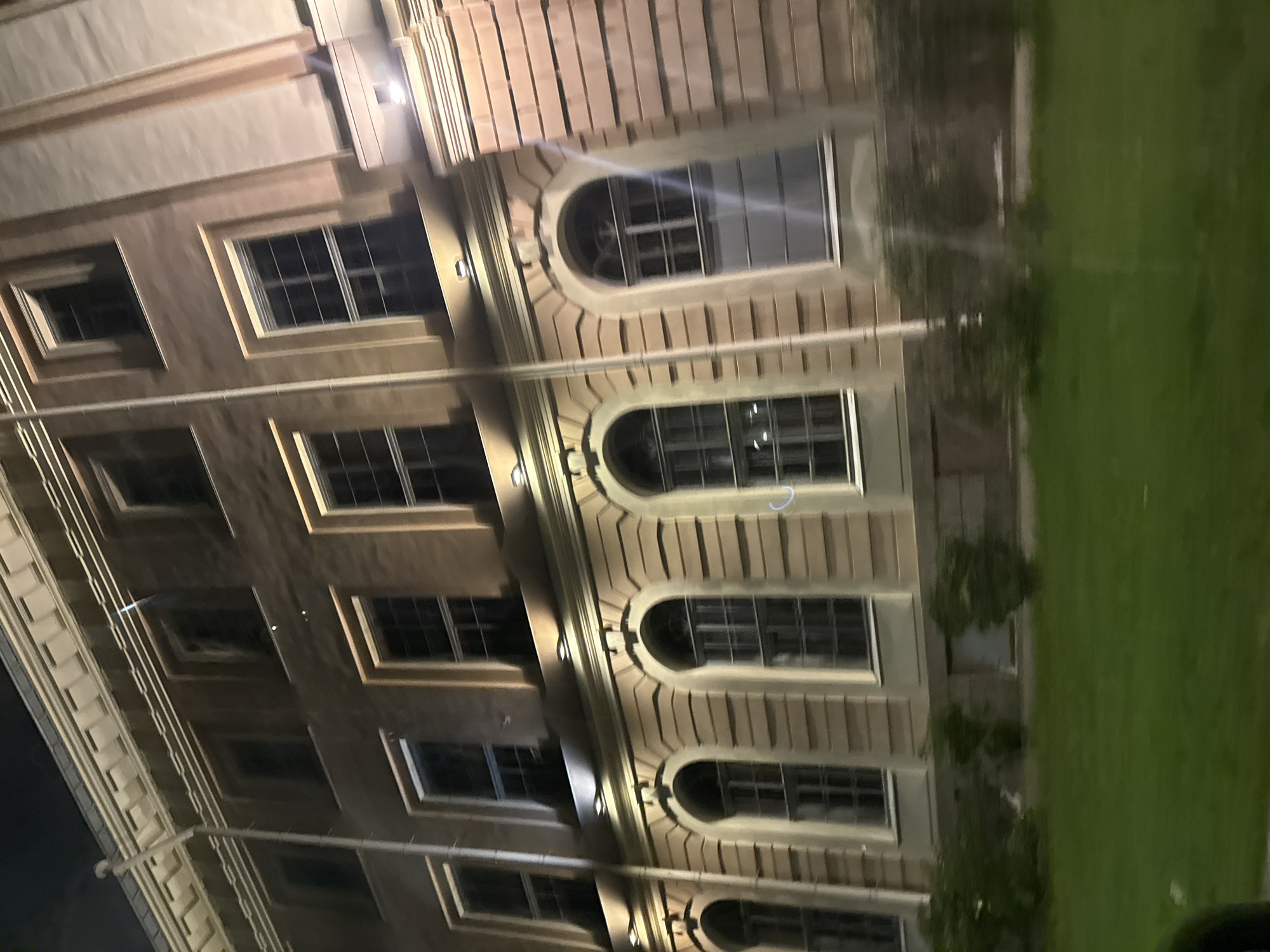 The image shows a multi-story building in Saint Petersburg, Russia, likely at night due to the lighting. The building has a light-colored facade with decorative elements around the windows and along the roofline. There are several rows of windows, some rectangular and some arched, many of which appear dark, suggesting they are not illuminated from within. Outdoor lights are visible illuminating the facade, creating highlights and shadows. In front of the building, there is a strip of green lawn and some dark bushes or trees. The overall impression is of an architectural structure with a formal design, possibly a public building or a historic residence. The lighting and shadows suggest it is dusk or nighttime.