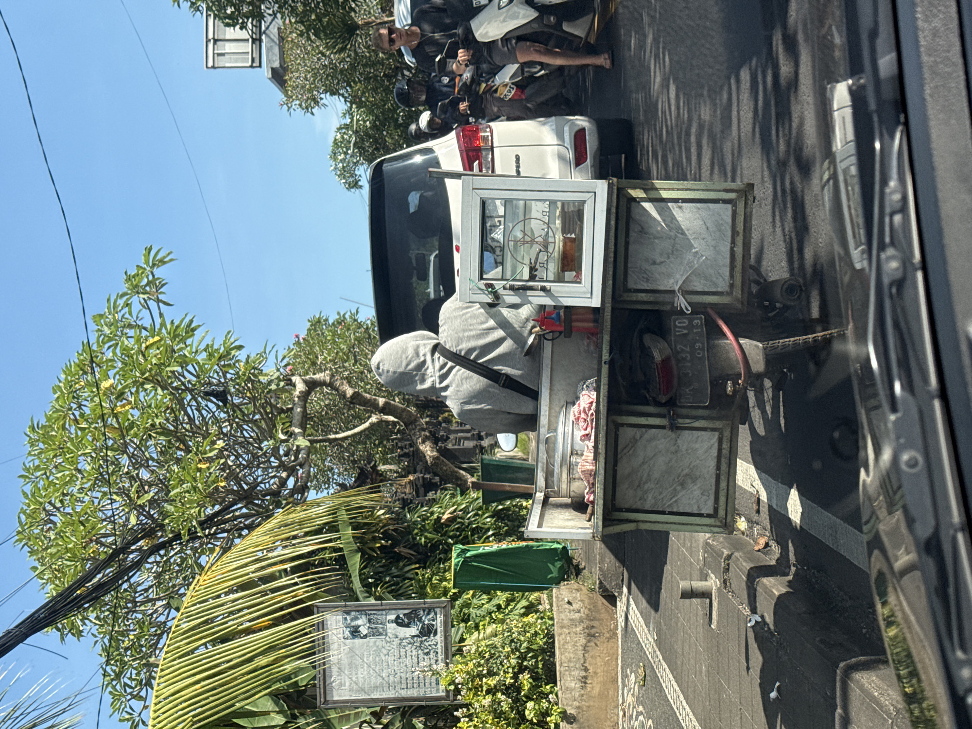 A street scene in Pecatu, Indonesia, shows a mobile food cart attached to a motorcycle parked behind a white SUV in traffic. The vendor, wearing a grey hooded sweatshirt, stands behind the cart, which has marble-patterned panels and an open display window. Other motorbikes with passengers are visible behind the SUV. Lush greenery and palm trees are present along the roadside. A signpost with a framed informational display stands amidst the foliage. The photo is taken from inside a vehicle, with its dashboard partially visible.