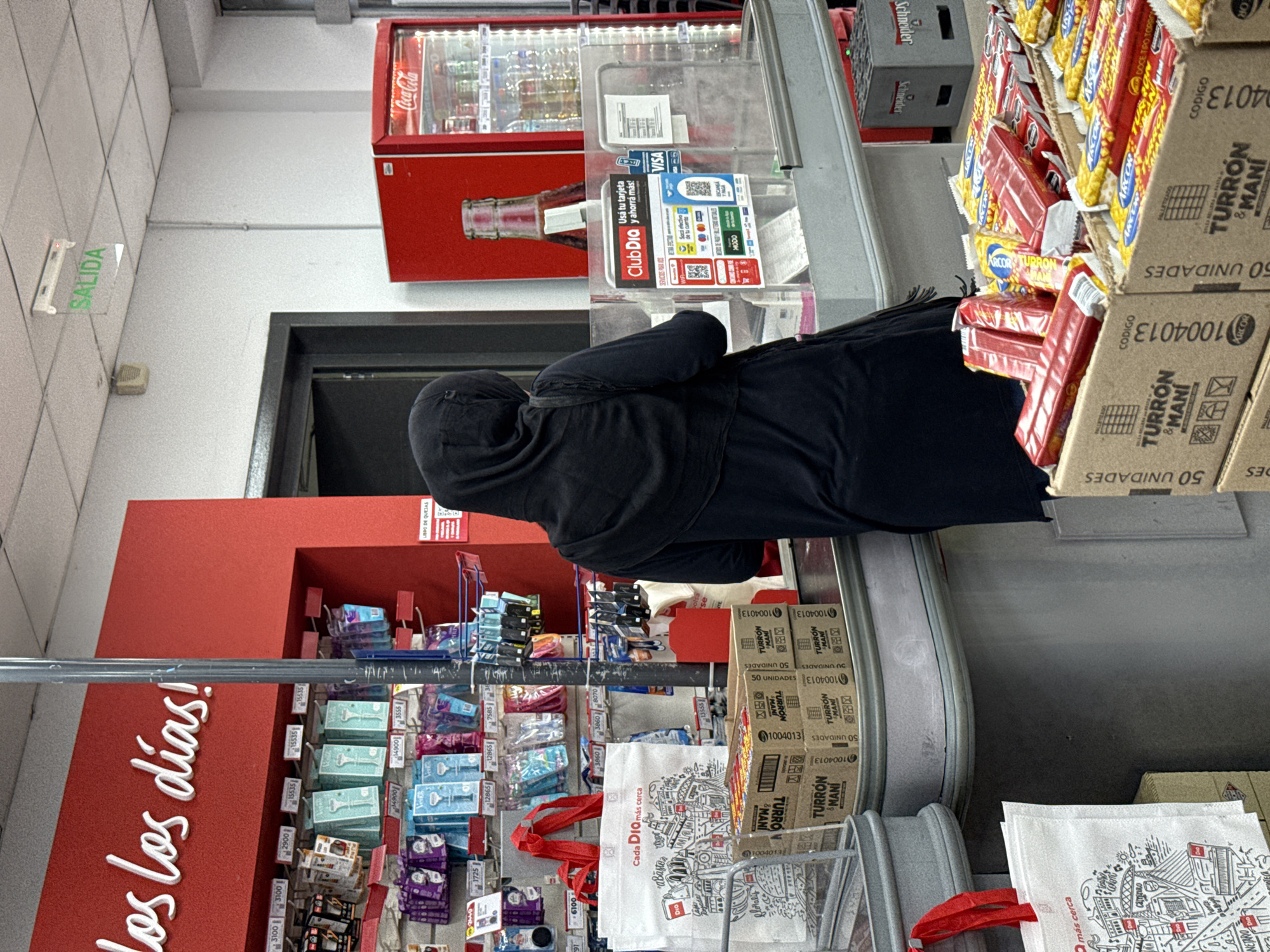 The image shows the interior of a Día supermarket, likely in Buenos Aires, Argentina, given the "Club Día" signage and promotional materials in Spanish.
A person wearing a dark, hooded jacket is standing at the checkout counter. Various products are visible in the foreground, including packaged sweets (turrón), razor blades, and reusable shopping bags. A Coca-Cola refrigerator unit stands behind the checkout area. The checkout counter displays signs promoting the Club Día loyalty program and indicating that Visa cards are accepted. The supermarket's branding is visible on a large red wall with the slogan "Todos los días!" (Every day!). The overall impression is a typical retail scene in a supermarket setting.