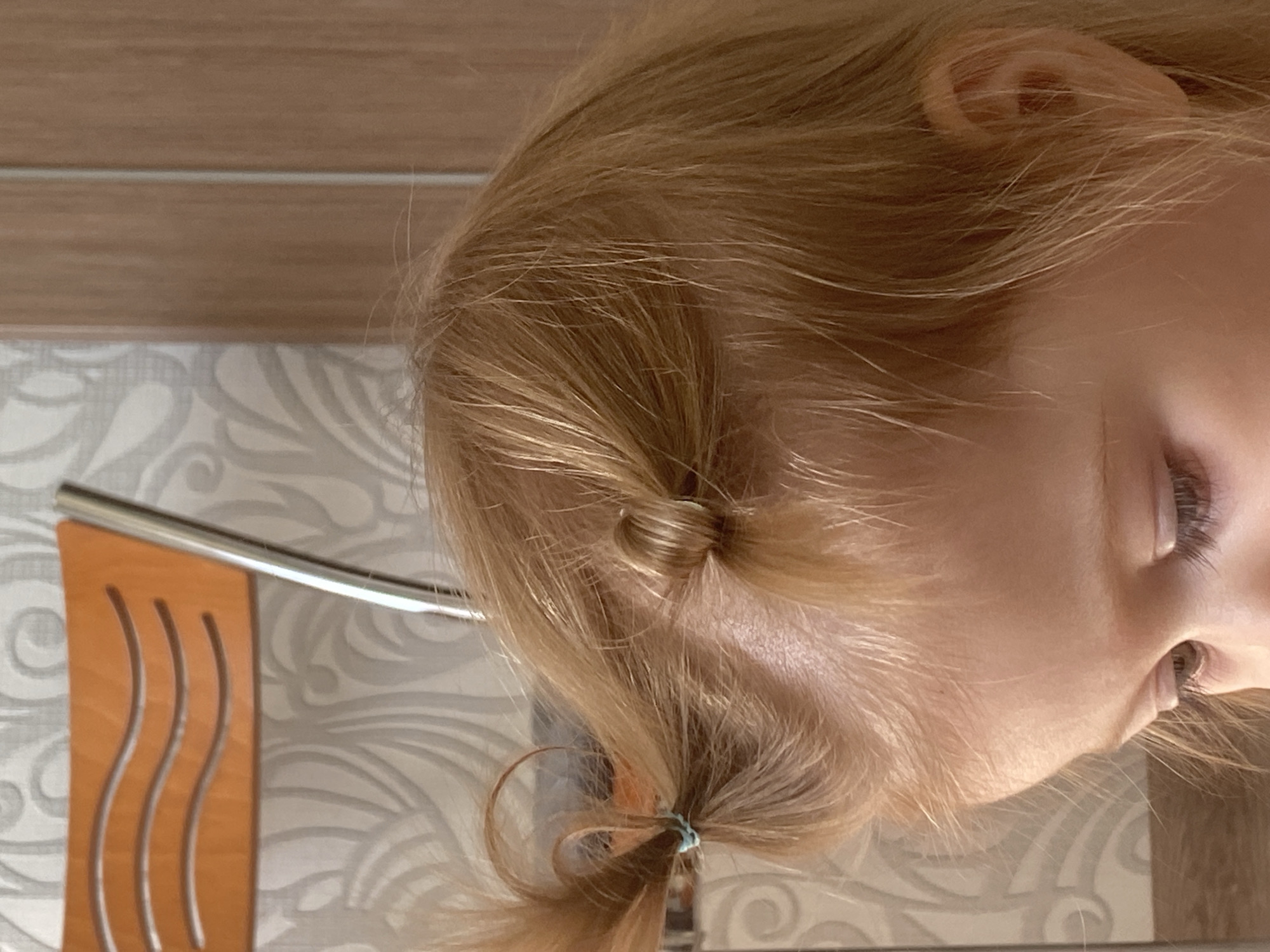 The image shows a close-up view of a young child's head, specifically the top and side. The child has blonde hair styled into two small pigtails, each secured with a light blue hair tie. The hair is fine and appears to be light brown or strawberry blonde. The child's skin is fair. In the background, a portion of an orange chair with a curved, slatted design is visible to the left, and a wooden-grained surface, likely a door or wall panel, is on the right. A patterned wallpaper with a swirling, abstract design is behind the chair. The lighting suggests an indoor setting. The specific location is Unknown, Unknown.
