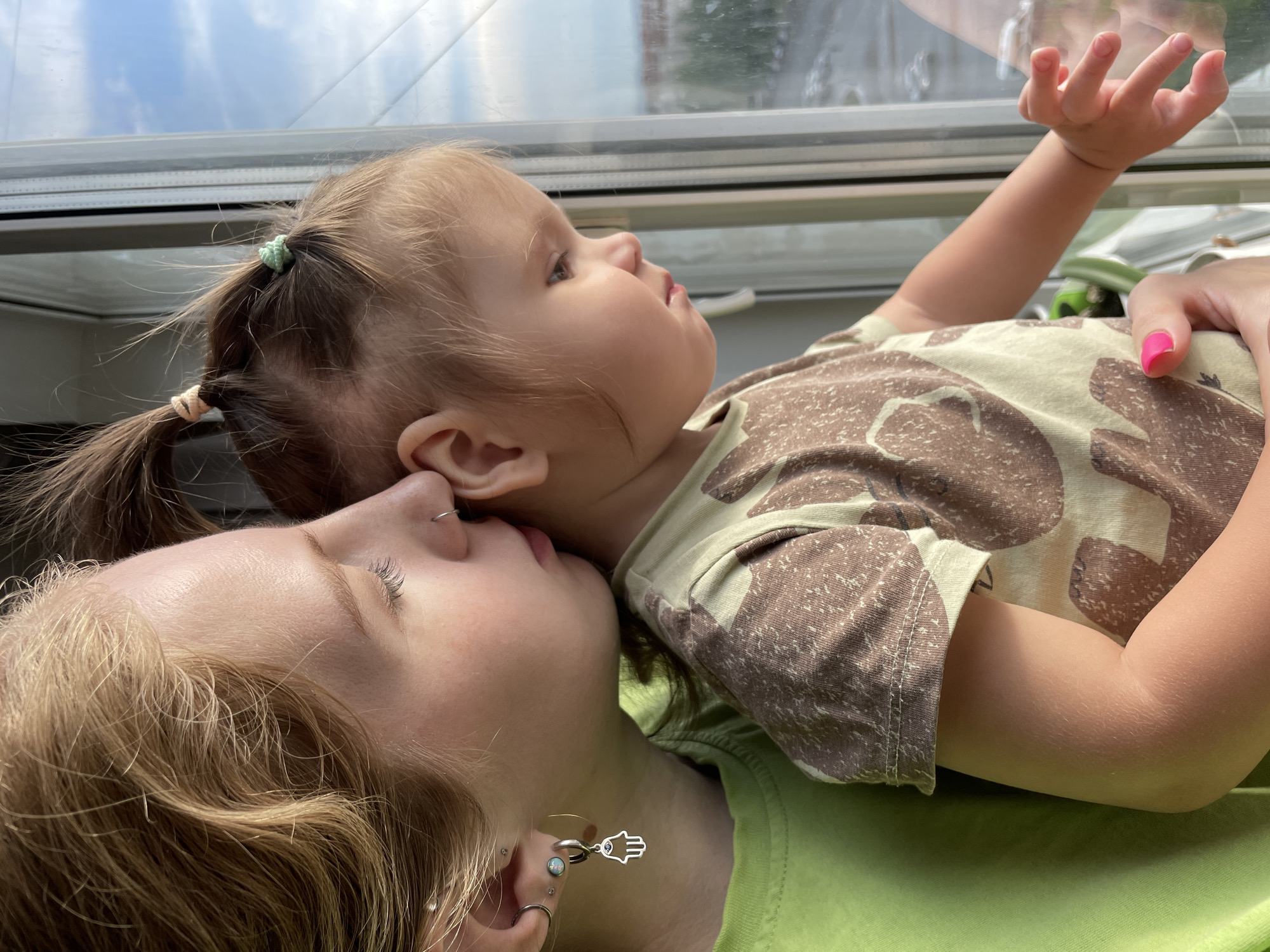 An intimate, high-angle shot captures two individuals, likely an older child or adult and a toddler, resting together near a window. The older individual is in the foreground, with light blonde hair falling over their face, a silver hoop nose piercing, and a distinctive Hamsa hand earring on their left ear. They wear a bright green top and appear to be lying down, possibly with eyes closed or partially open, facing away from the viewer.
Resting on the older person's shoulder or chest is a young child or toddler, with light brown hair styled in two small ponytails, secured with a light green and a beige scrunchie. The child wears a light brown and off-white camouflage-patterned t-shirt. Their face is in profile, looking upwards and outwards through the window, with one small hand raised towards the glass as if reaching for something outside. The older person's hand, with bright pink nail polish, is gently resting near the child's body.
The background shows a bright, somewhat overcast sky with clouds reflected in the windowpanes, indicating daytime. The scene evokes a sense of tenderness, curiosity, and quiet observation. The setting appears to be an indoor space, possibly a home, in Izhevsk, Russia. No visible text is present in the image.