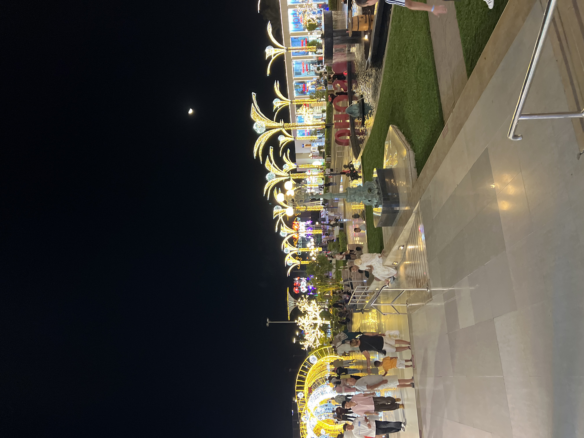The image shows a nighttime scene at a decorated outdoor public space in Sharm El Sheikh, Egypt. The area is illuminated with abundant string lights and decorative lighting fixtures resembling palm trees. People are seen walking along a paved walkway, some in groups and some individually. In the background, there are shops and commercial establishments, also decorated with lights. A fountain is visible with a water feature. A crescent moon is faintly visible in the dark sky. The overall atmosphere suggests a festive or recreational setting.