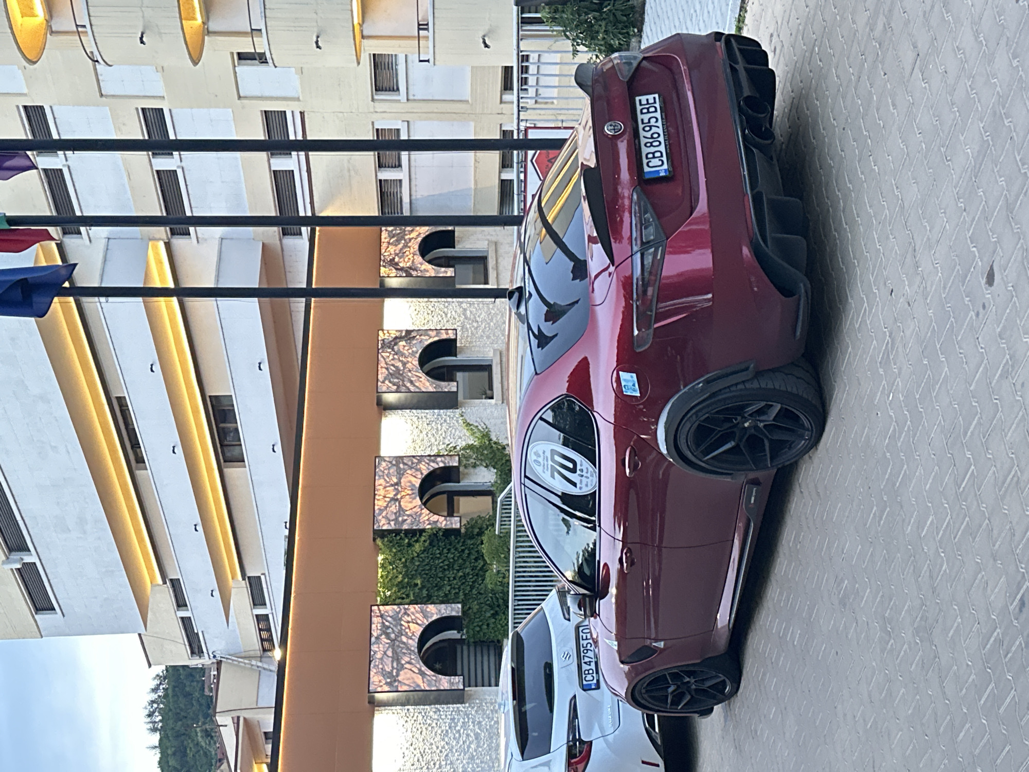 The image shows a red Alfa Romeo Giulia parked on a paved surface outside a modern building in Veliko Tarnovo, Bulgaria. The car has a spoiler and a diffuser, and black rims. A white Suzuki Swift is parked to the left of the Alfa Romeo. The building behind the cars has multiple balconies with orange lighting. Several flags are visible on poles in front of the building. The sky is overcast.