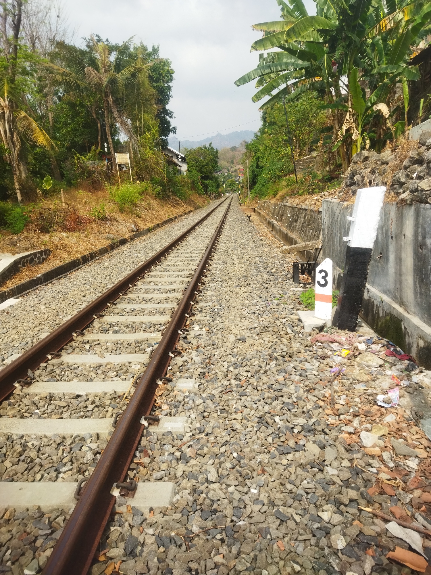 The image depicts a pair of railroad tracks in Giripurwo, Indonesia, receding into the distance towards rolling hills under a cloudy sky. The tracks are laid on a ballast of gray and brown gravel, with concrete sleepers visible beneath the steel rails. To the right of the tracks, a concrete wall lines the embankment, which is overgrown with dry grass and some rocks. At the base of this wall, a white marker post with a black and orange stripe and the number "3" is planted. Scattered debris, including plastic bags and fabric scraps, lies around the marker post. A steep, vegetated hillside rises to the left of the tracks, with various trees, including palm trees, and some structures visible amongst the foliage. In the background, the landscape transitions to hills covered in vegetation. The scene is static and shows no people or discernible activity.
