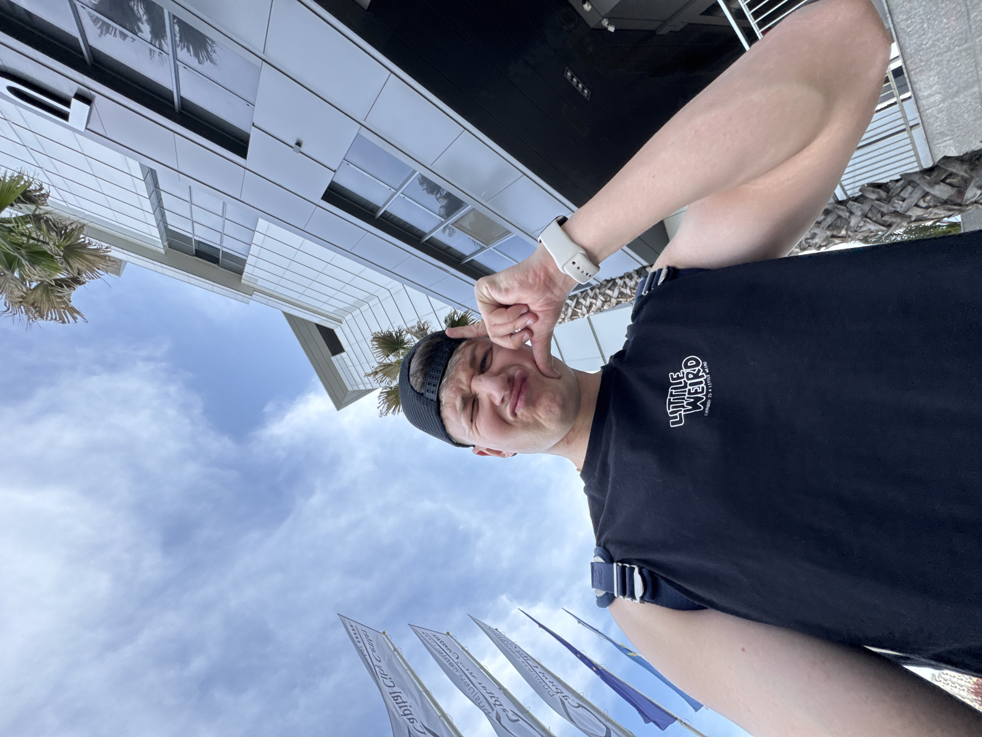 The image, taken from a low-angle perspective and rotated 90 degrees counter-clockwise from its upright orientation, features a young man striking a playful pose against a backdrop of modern buildings and an overcast sky.
When viewed upright, the man is centered, looking upwards and to his right with a pouting, scrunched-up facial expression. His left hand is raised near his face, forming an "L" shape with his thumb and index finger. He is wearing a black short-sleeved t-shirt with the text "LITTLE WEIRD" and "EVERYONE IS A LITTLE WEIRD" printed on the chest. A black headband or cap covers his hair, and he has a white smartwatch on his left wrist, along with a dark strap from a bag or camera over his right shoulder.
To the man's left (frame left when upright), modern buildings with white and grey paneling and large, reflective windows rise into the sky. Reflections of palm trees are visible in some windows, and actual palm foliage can be seen near the buildings. The upper part of the frame is filled with a bright, partly cloudy or overcast sky. In the lower left foreground, several tall, narrow flags are visible. Most are white with dark text, while one appears blue/purple. The text on the white flags, partially obscured and reversed, reads "Plovdiv European Capital of Culture," confirming the location context of Plovdiv, Bulgaria.
The scene depicts an outdoor setting during daytime under cloudy conditions. The man's pose and the unique camera angle suggest a playful selfie.