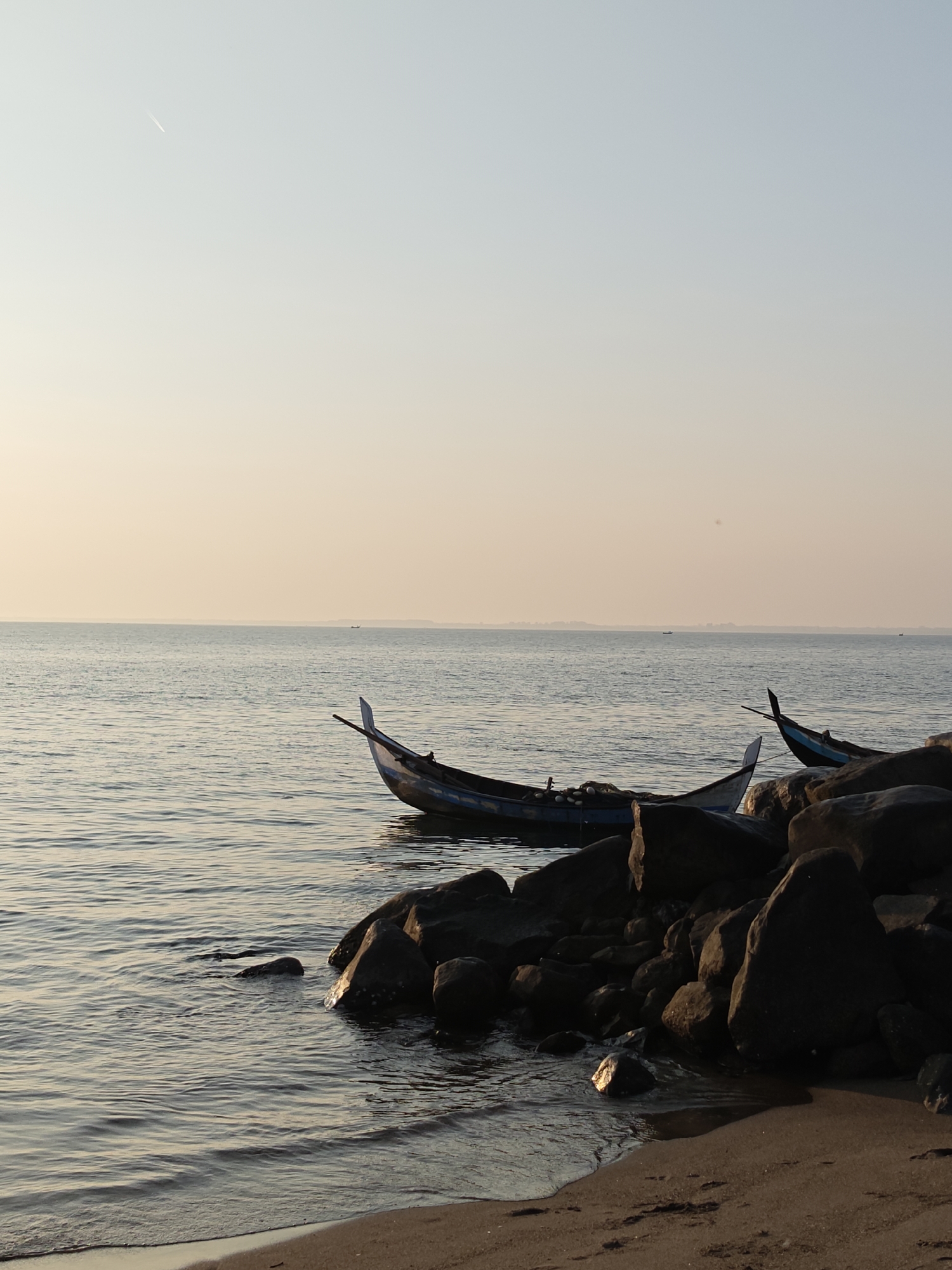 The image shows a coastal scene in Lhokseumawe, Indonesia, at what appears to be dawn or dusk, given the soft, warm light. Two traditional fishing boats are visible; one is beached against a collection of dark rocks near the shoreline, and the other is partially visible to the right. The sea is calm with gentle ripples and reflections of the sky. The sky is a pale gradient of light blue and orange. The beach is sandy and slopes slightly into the water. An aircraft contrail can be seen in the upper left portion of the sky. In the distance, the horizon is visible with outlines of land.