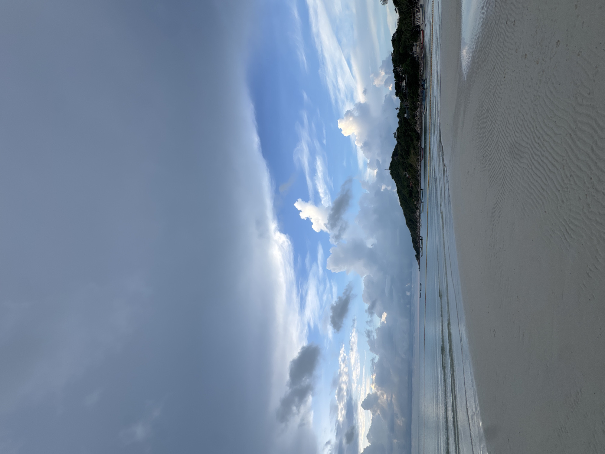 The image shows a tranquil beach scene at what appears to be dusk in Ban Tai Subdistrict, Thailand. The foreground is dominated by a wide expanse of pale sand with distinct ripple patterns created by receding water. The tide is low, revealing wet sand that reflects the overcast sky.
In the mid-ground, the calm, shallow water of the sea gently laps the shore. Several small, traditional Thai fishing boats are moored or anchored near the coastline. A yellow floating rope barrier stretches across a portion of the water, delineating an area.
The background features a lush, green, hilly island covered with dense vegetation and scattered buildings. A prominent structure, possibly a lighthouse or a tower, is visible on the highest point of the hill. The island is fringed with more buildings and palm trees along its base.
The sky above is a dramatic display of clouds. The upper portion is filled with heavy, dark gray storm clouds, suggesting recent or approaching rain. Below these, the clouds break up into patches of white cumulus and wispy cirrus clouds, interspersed with patches of bright blue sky, hinting at a transition in the weather. The overall lighting is diffused and soft, characteristic of a cloudy evening. No people are visible in the frame.