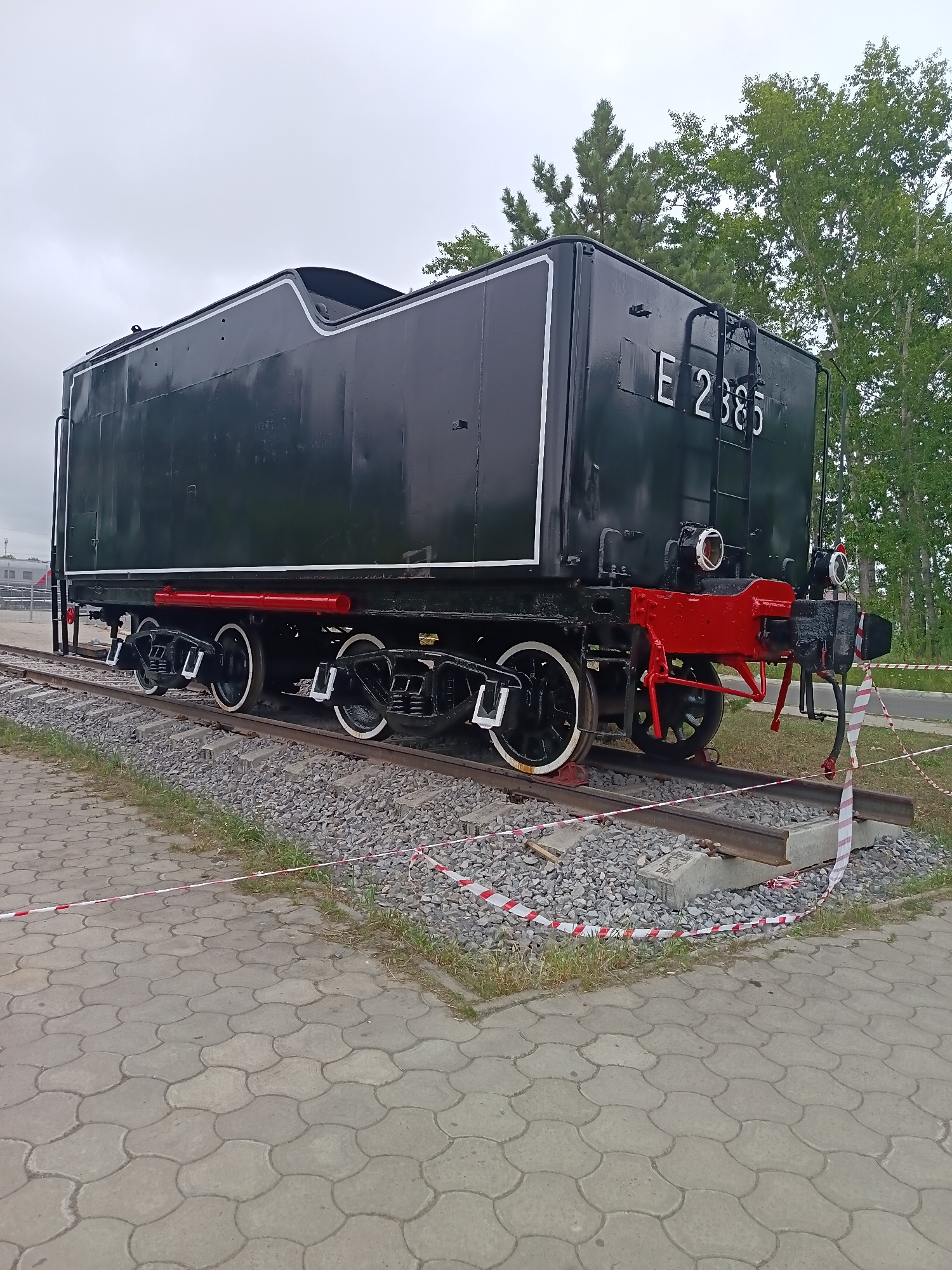 The image shows a black steam locomotive, identified by the marking "E 2385," positioned on railway tracks in Komsomolsk-on-Amur, Russia. The locomotive is dark colored with white and red accents, and its wheels are visible. The scene is outdoors, with a cloudy sky overhead and trees in the background. The tracks are surrounded by gravel and a concrete pathway is visible in the foreground, demarcated by red and white caution tape. The locomotive appears to be a static exhibit, with no people visible in the frame. The setting suggests an outdoor display area, possibly a museum or a historical site within the city.