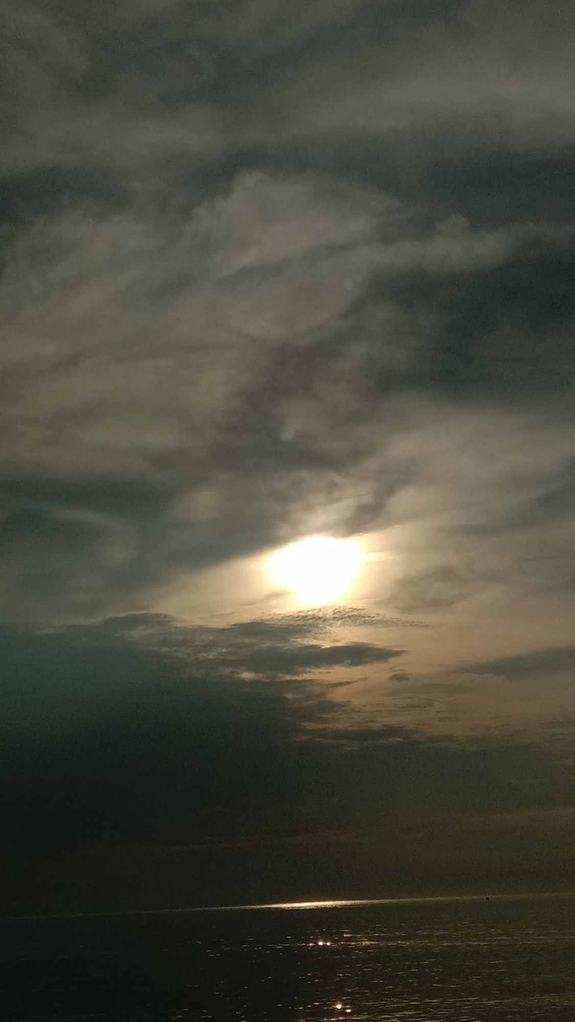 The image depicts a dark and cloudy sky over the sea, likely taken at dusk or dawn. A bright light, possibly the sun, shines through a gap in the clouds, creating a path of reflected light on the water's surface. The sky is filled with grey and white clouds. The water appears calm with gentle ripples. No people or specific objects are visible. The location could be Lhokseumawe, Indonesia, given the request, but this cannot be verified from the image alone.