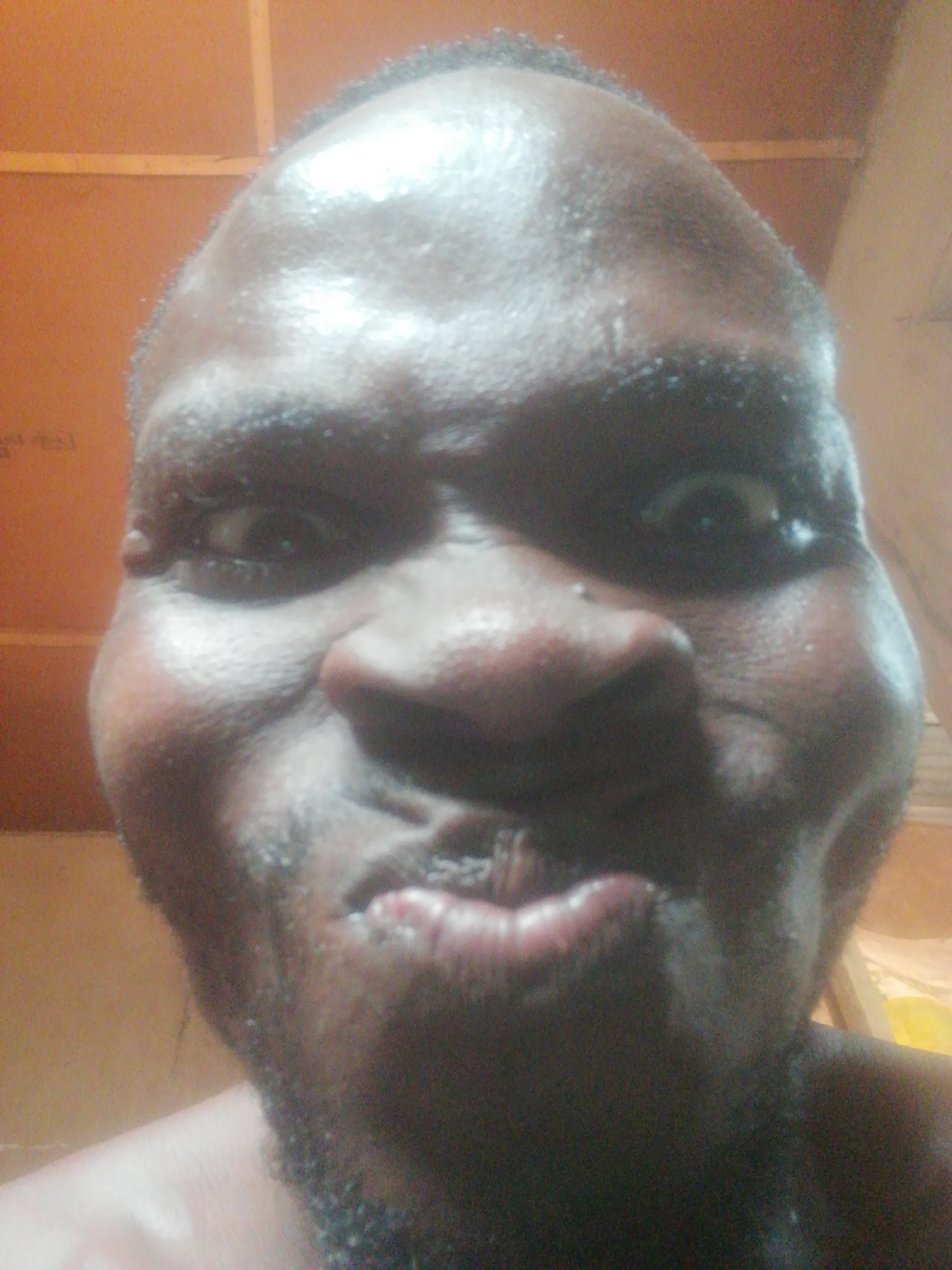 A close-up selfie of a dark-skinned male with a shaved head. He has a wide nose and full lips, and his cheeks are puffed out. He has dark eyebrows, and his eyes are slightly wide. He has some stubble on his chin. The background is out of focus and appears to be an interior space with wood paneling. The location is Unknown, Unknown.
