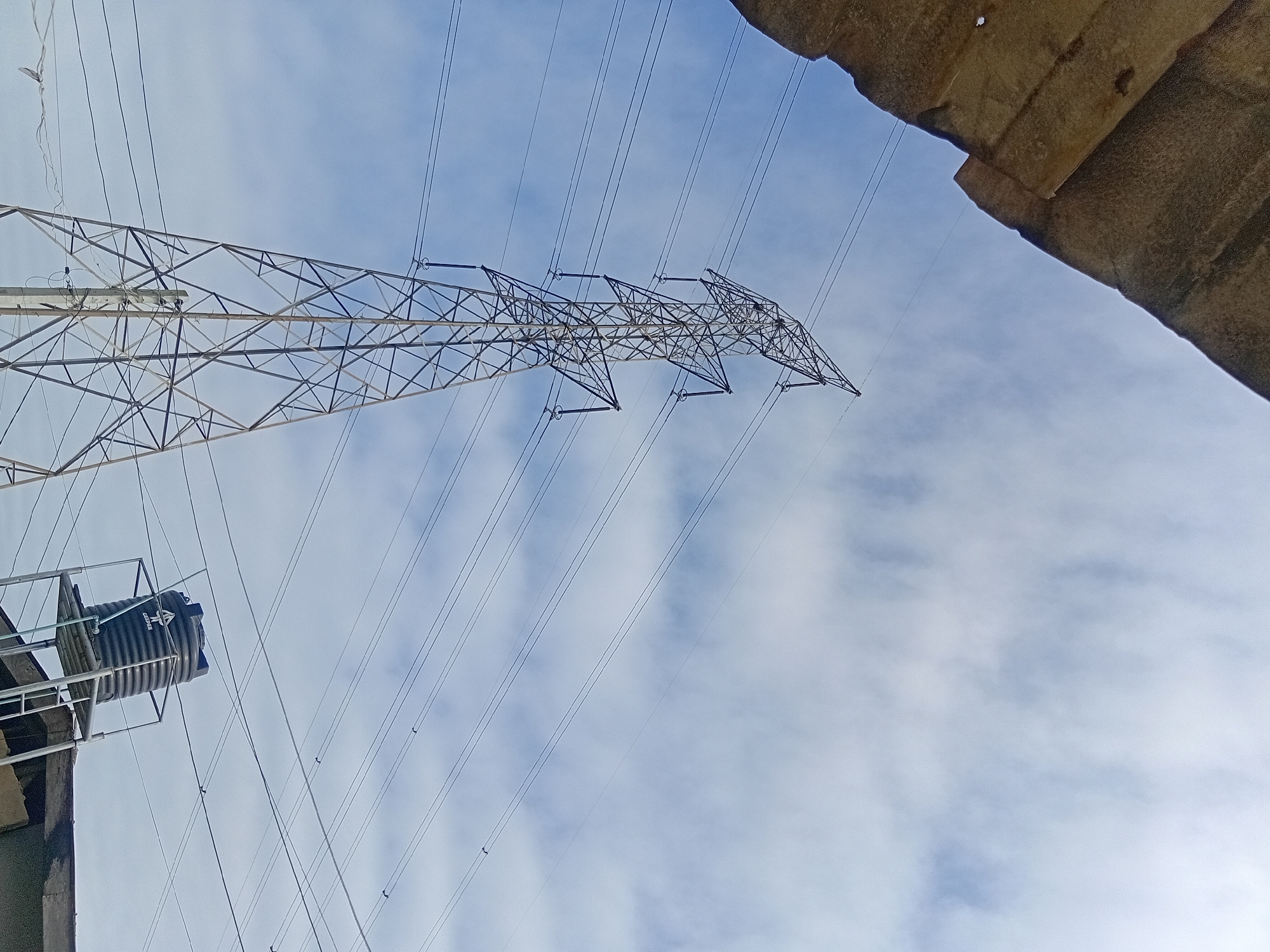 A high-angle, wide shot captures a tall, lattice-style electrical tower against a partly cloudy sky. The tower, situated in an unspecified urban location, is a prominent feature, with several thick, black power lines extending from it and disappearing into the upper right quadrant of the frame. In the lower right, a dark, ribbed water tank sits atop a silver metal platform, which is supported by a ladder. The top left of the image is partially obscured by a rough, textured, brown structure, possibly a roof or overhang. The sky is a pale blue, with scattered white clouds that have a wispy appearance.