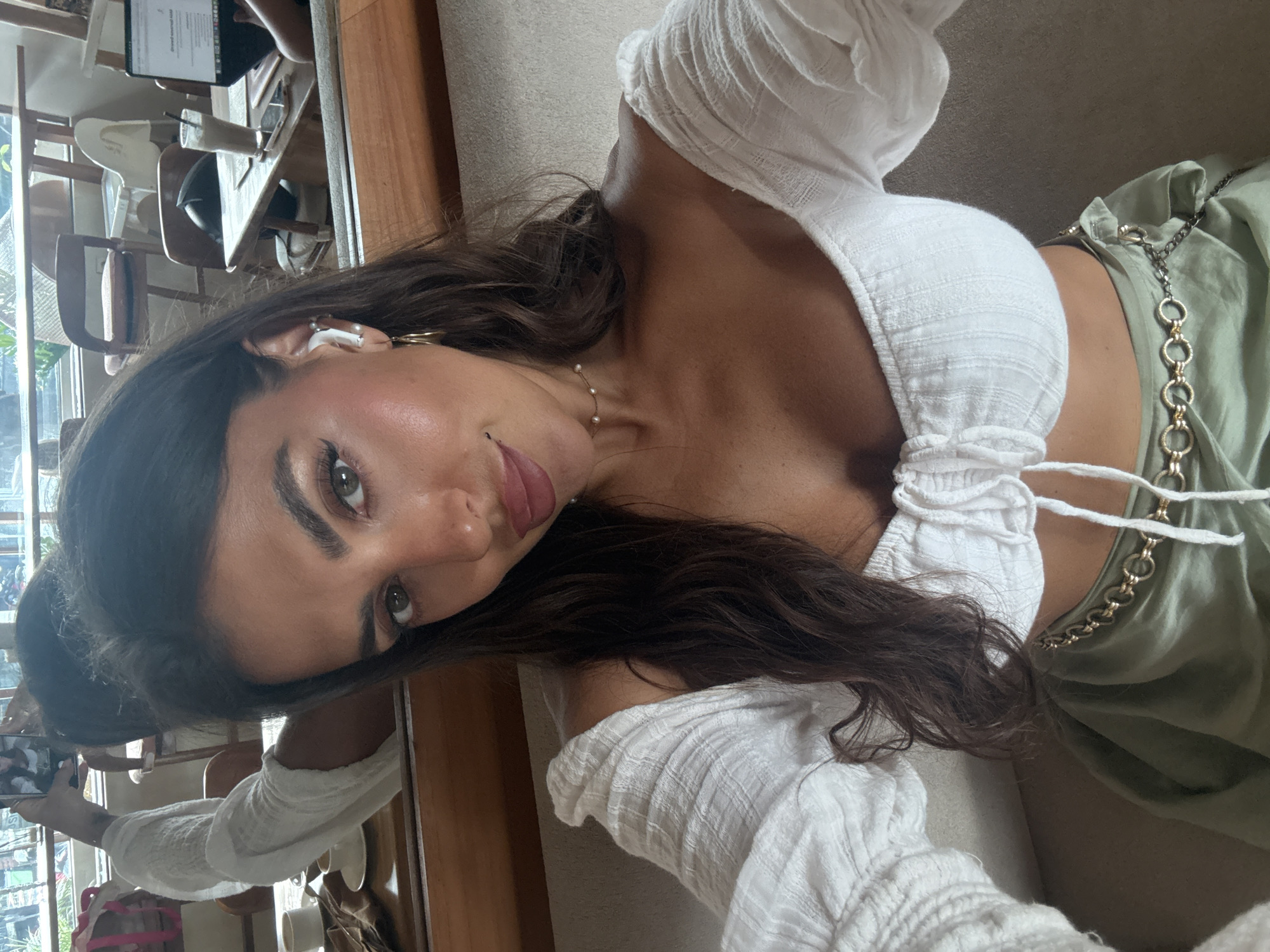 The image depicts a woman in what appears to be an indoor setting, possibly a café or restaurant. She is holding a cell phone and taking a selfie. The woman has long, dark hair and is wearing a white, textured top and olive-green shorts with a decorative gold chain belt. An earbud is visible in her left ear. In the background, there is a partial view of a table with a tablet, chairs, and outdoor scenery through a window or opening, hinting at a well-lit, open-air establishment. The image does not confirm that the location is North Kuta, Indonesia, as it is not possible to determine the exact location from the provided image.