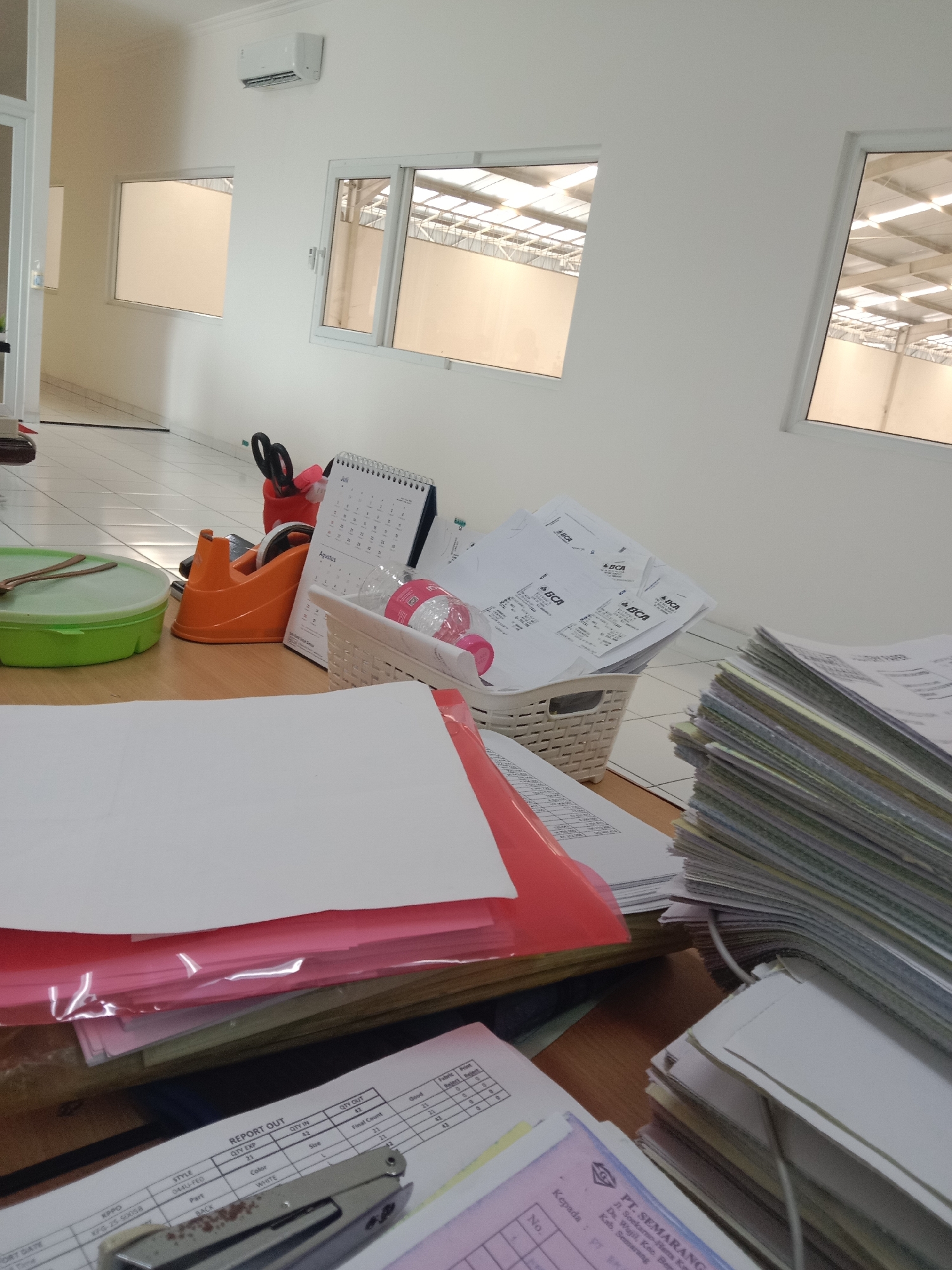 The image shows a cluttered office desk in what appears to be a warehouse or industrial space. The room has white walls, a tiled floor, and large windows looking out onto a structure with a translucent roof, suggesting a commercial or manufacturing environment. On the desk, there are stacks of papers, a stapler, a tape dispenser, a water bottle, scissors, a green container, and a calendar. One document on the desk has the logo "PT. SEMARANG" and an address that places the location in Semarang, Indonesia. The overall impression is one of a busy, functional workspace.