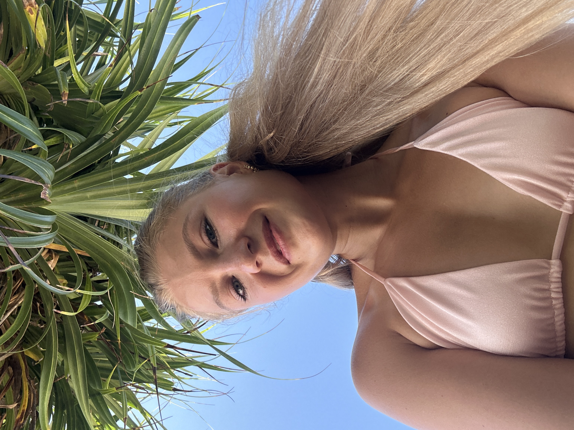 A woman in a light pink bikini stands near a palm tree, looking down at the camera with a slight smile. The clear blue sky is visible in the background. The location is likely in Kuta Selatan, Indonesia, given the presence of the palm tree and the woman's attire, suggesting a beach setting.