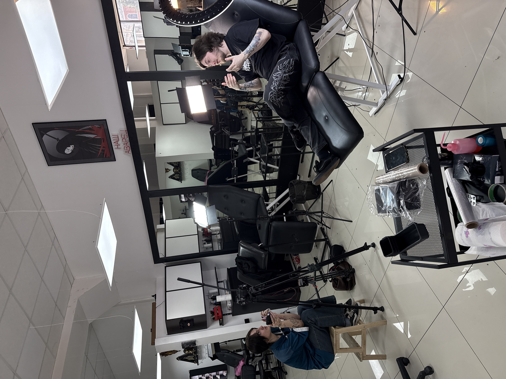 The photograph shows the interior of a tattoo studio in Rostov-on-Don, Russia. One man sits in a black tattoo chair, wearing a black T-shirt, patterned pants, and sneakers, and has visible tattoos on his arms. He appears to be talking with his hands. Another man sits on a wooden stool in front of video equipment, wearing a blue striped shirt and jeans. He also has tattoos on his arms and is holding a phone. Various pieces of tattoo equipment, including lights, tripods, and a metal storage rack, are visible. A framed picture is on the wall, as is what appears to be a sign written in Russian that says "наши аварели". The floor is tiled in white. A large mirror reflects the scene.