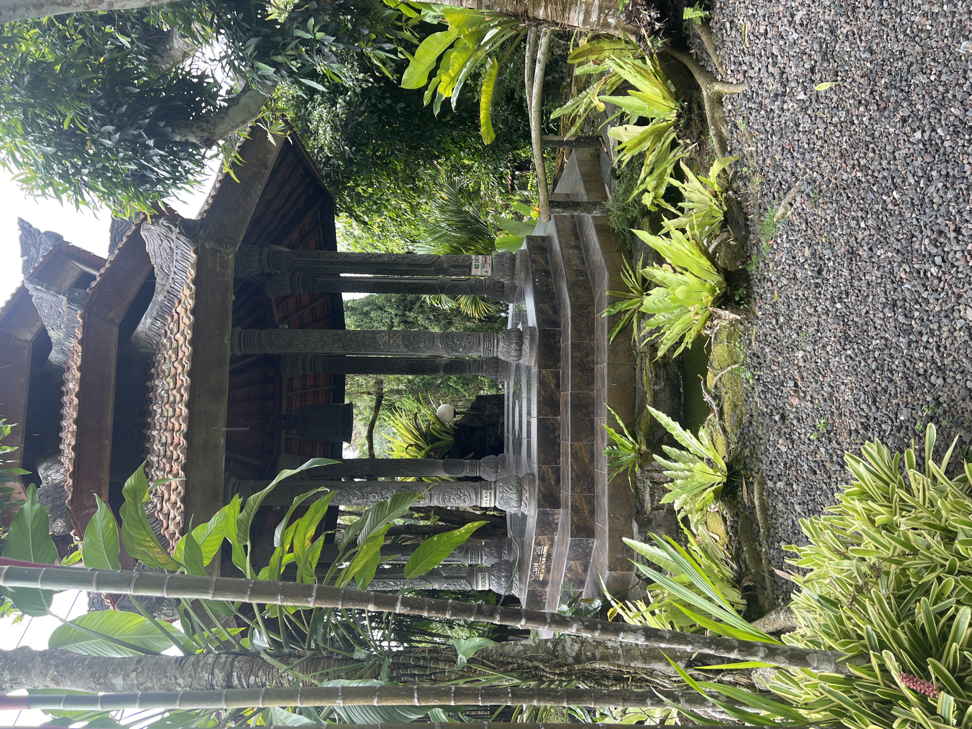 The image depicts a traditional Balinese gazebo situated in a lush, verdant garden environment in Pupuan, Indonesia. The gazebo features a multi-tiered, brown tiled roof supported by intricately carved stone pillars, some of which bear inscriptions and decorative motifs. A set of tiered stone steps leads up to the central platform of the gazebo. Surrounding the gazebo are various types of tropical plants, including large, leafy ferns and tall bamboo stalks. The ground is covered with dark grey gravel, and a small pond or water feature is visible near the base of the gazebo. A rustic wooden railing is present to the right of the steps. The overall scene conveys a sense of tranquility and natural beauty characteristic of a tropical garden setting. There are no people visible in the image.