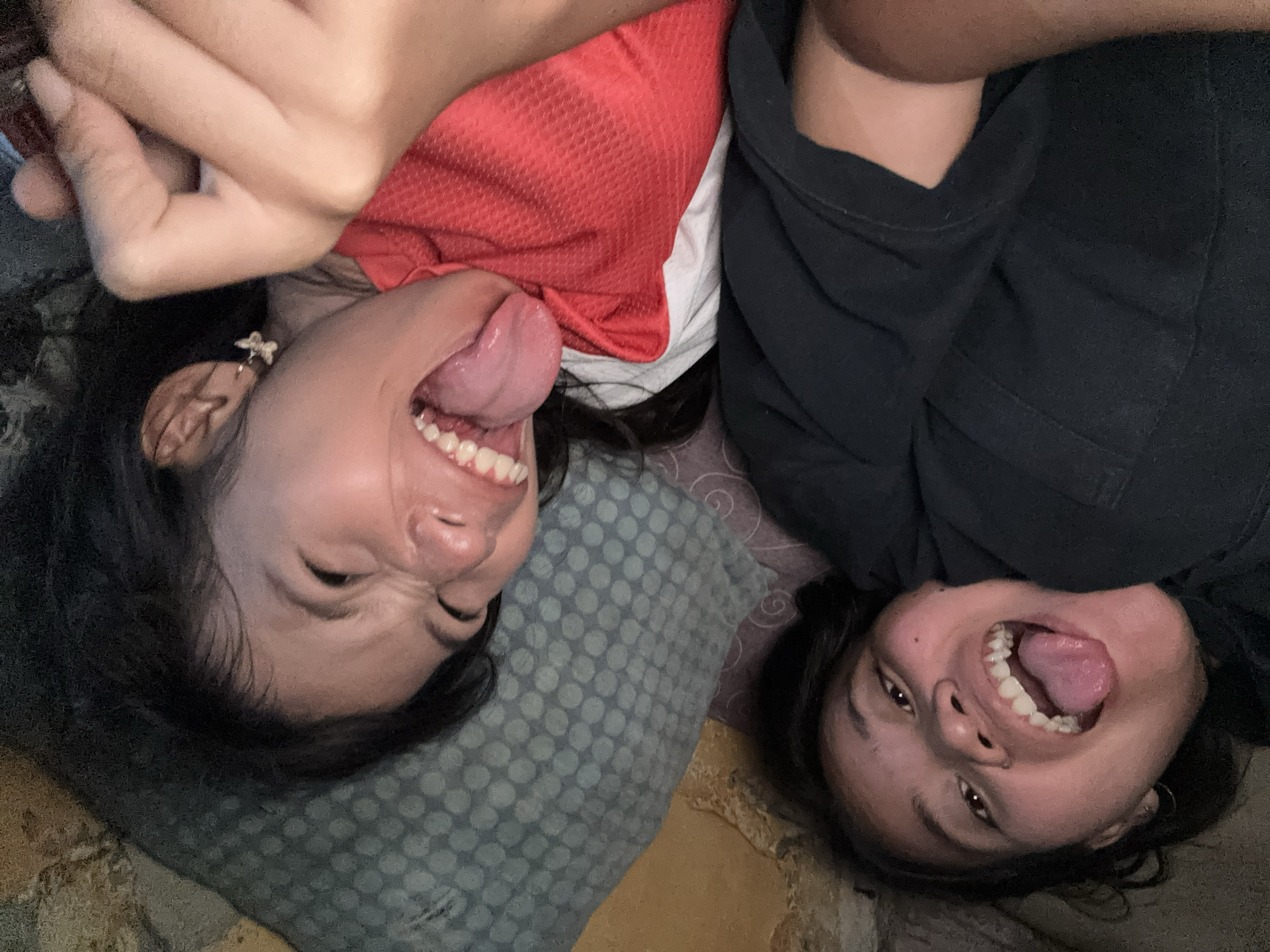 The media file is a close-up, upside-down selfie of two young women lying down, with their faces angled towards the camera. They are on a bed, suggested by the pillows and bedding visible. One woman is wearing a red top, and the other is wearing a black top. Both women have their mouths wide open, with their tongues sticking out, and appear to be making faces for the photo. They have dark hair and are wearing earrings. The lighting suggests it is indoors, possibly in the evening or a dimly lit room. The overall mood of the image is playful and lighthearted, as indicated by their exaggerated expressions. The location context is East Jakarta, Indonesia. No visible text is present in the image.