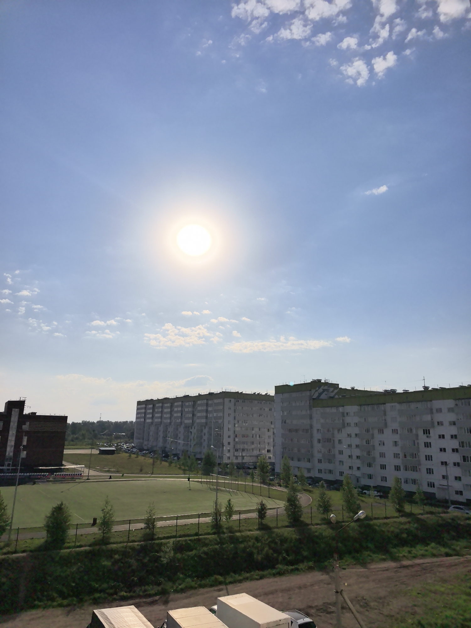The image depicts a sunny day in the city of Фролы, Russia. The sun is high in the sky, partially obscured by thin clouds. In the background, there are several large apartment buildings with a light gray facade and green roofs. In the middle ground, there is a fenced sports field with a green turf. Trees are planted along the fence line. In the foreground, the roofs of several parked trucks are visible. A dirt road is near the trucks.