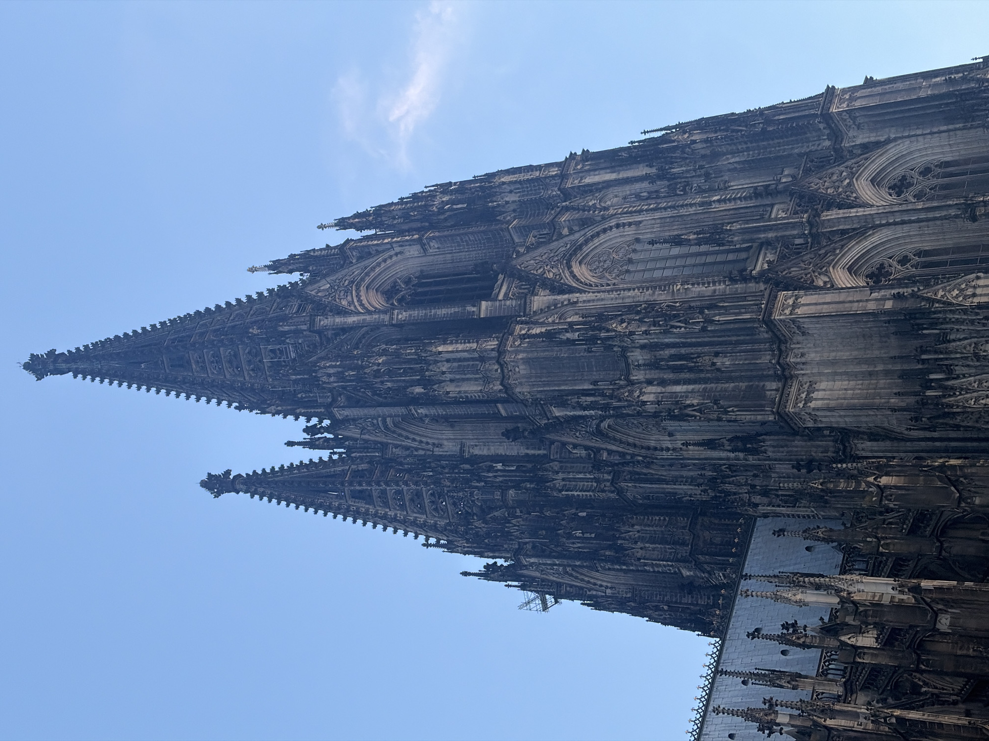The image shows a low-angle, close-up view of the Cologne Cathedral, also known as Kölner Dom, a Roman Catholic cathedral in Cologne, Germany. The towering Gothic architecture dominates the frame, with its intricate stonework, pointed spires, and arched windows. The sky is a clear, pale blue with a wispy cloud on the right side. The cathedral's dark, weathered stone contrasts with the bright sky. Part of the cathedral's roof, covered in grey shingles, is visible in the lower left portion of the image. Scaffolding is present on the upper left side of the cathedral, indicating ongoing restoration or maintenance work. No people are visible in the image.