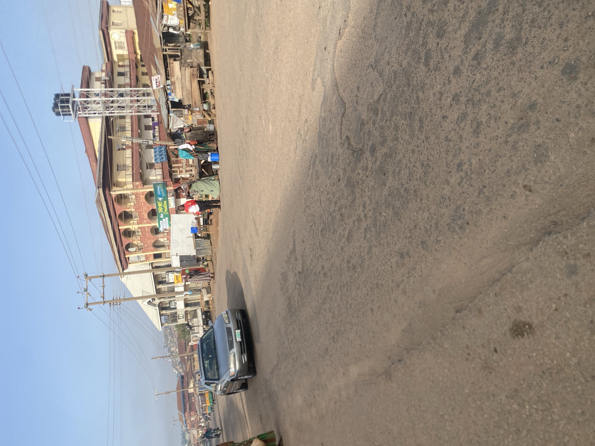 The image shows a street scene in Jos, Nigeria, under a clear blue sky. In the foreground, a grey sedan is driving on a paved road. The road surface is cracked and appears worn. To the right, there are market stalls with corrugated metal roofs and wooden structures. Several people are standing near the stalls, including a woman in a green dress and another in a red shirt and dark pants. In the background, there are multi-story buildings, one with a distinctive arched facade and another with a tall water tower. Utility poles and wires crisscross the sky. A few motorcycles and a motorized tricycle are visible further down the street, suggesting traffic. The overall impression is of a bustling urban environment with commercial activity.