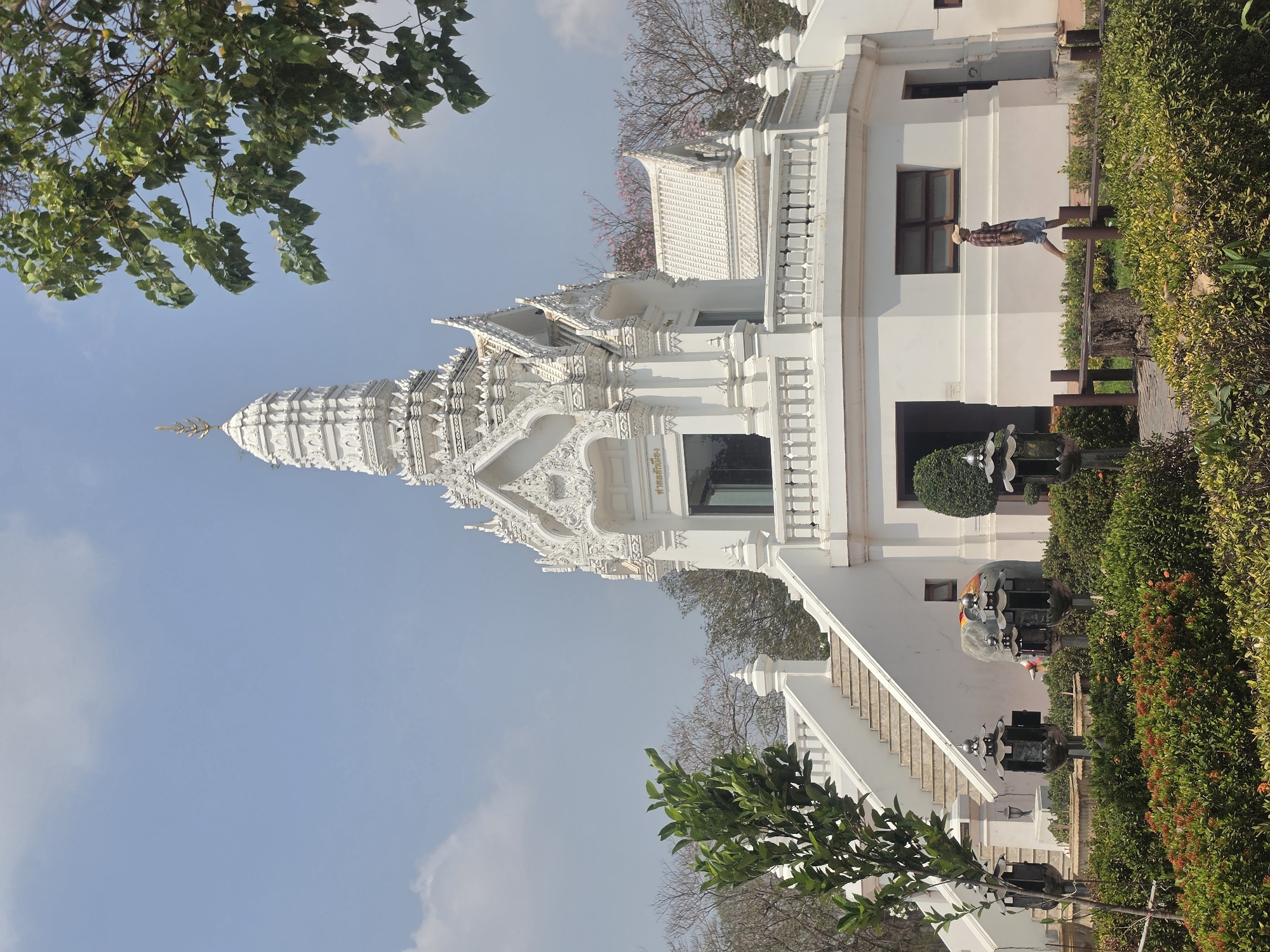 This image depicts Wat Ratchaburana, a Buddhist temple in the Historic City of Ayutthaya, Thailand. The temple is predominantly white, featuring intricate architectural details. A person stands on a bench, possibly observing the scene, next to the temple structure. Green vegetation borders the building, and the sky is partly cloudy.