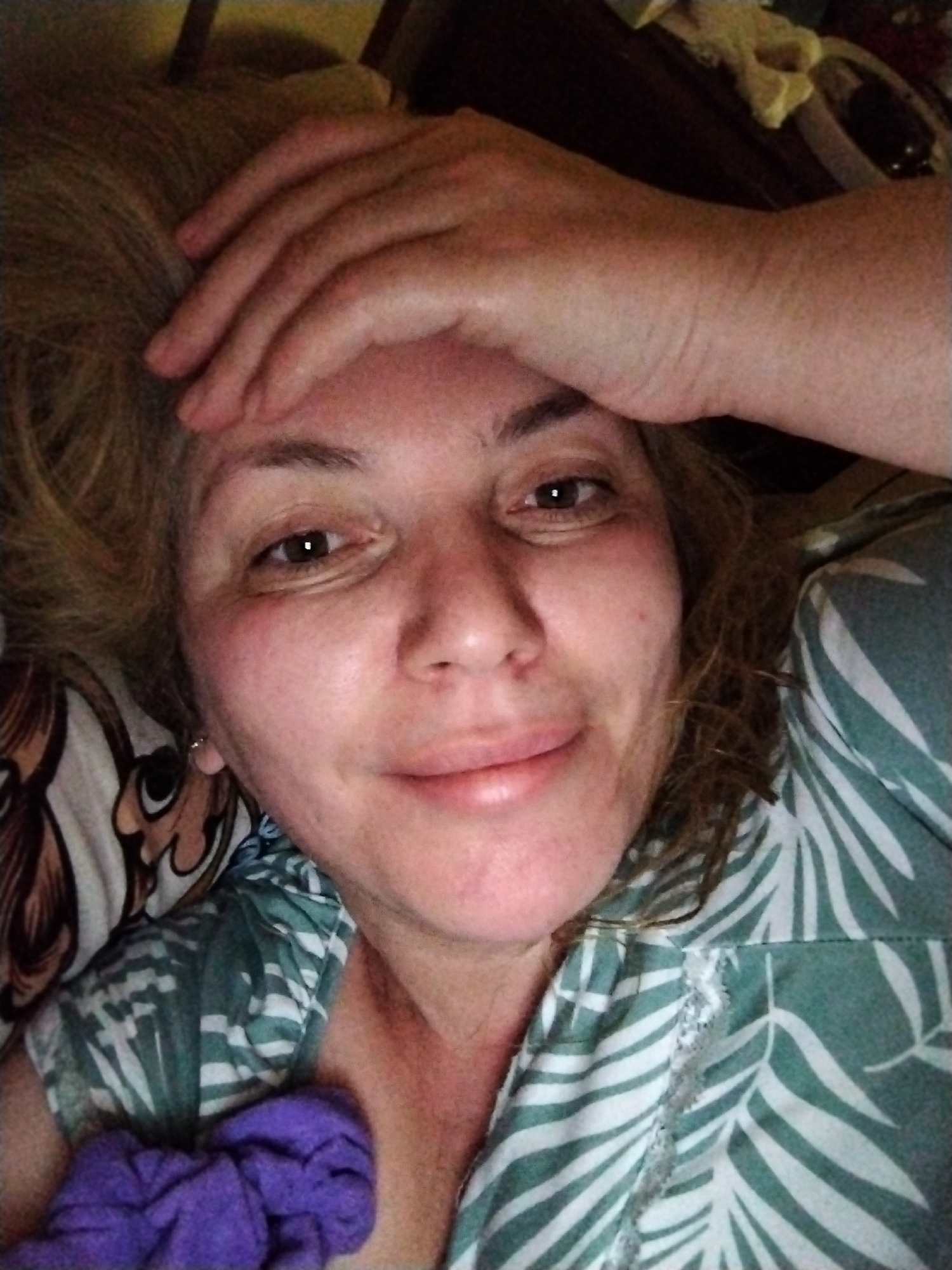 A woman is lying down, possibly in bed, as indicated by the patterned pillow and green and white palm leaf patterned shirt. She has light brown or blonde hair and is looking at the camera with a slight smile. Her hand is resting on her forehead. A purple object is visible near her chest. The location, Al Mas'oudi in the United Arab Emirates, is not discernible from the image itself.