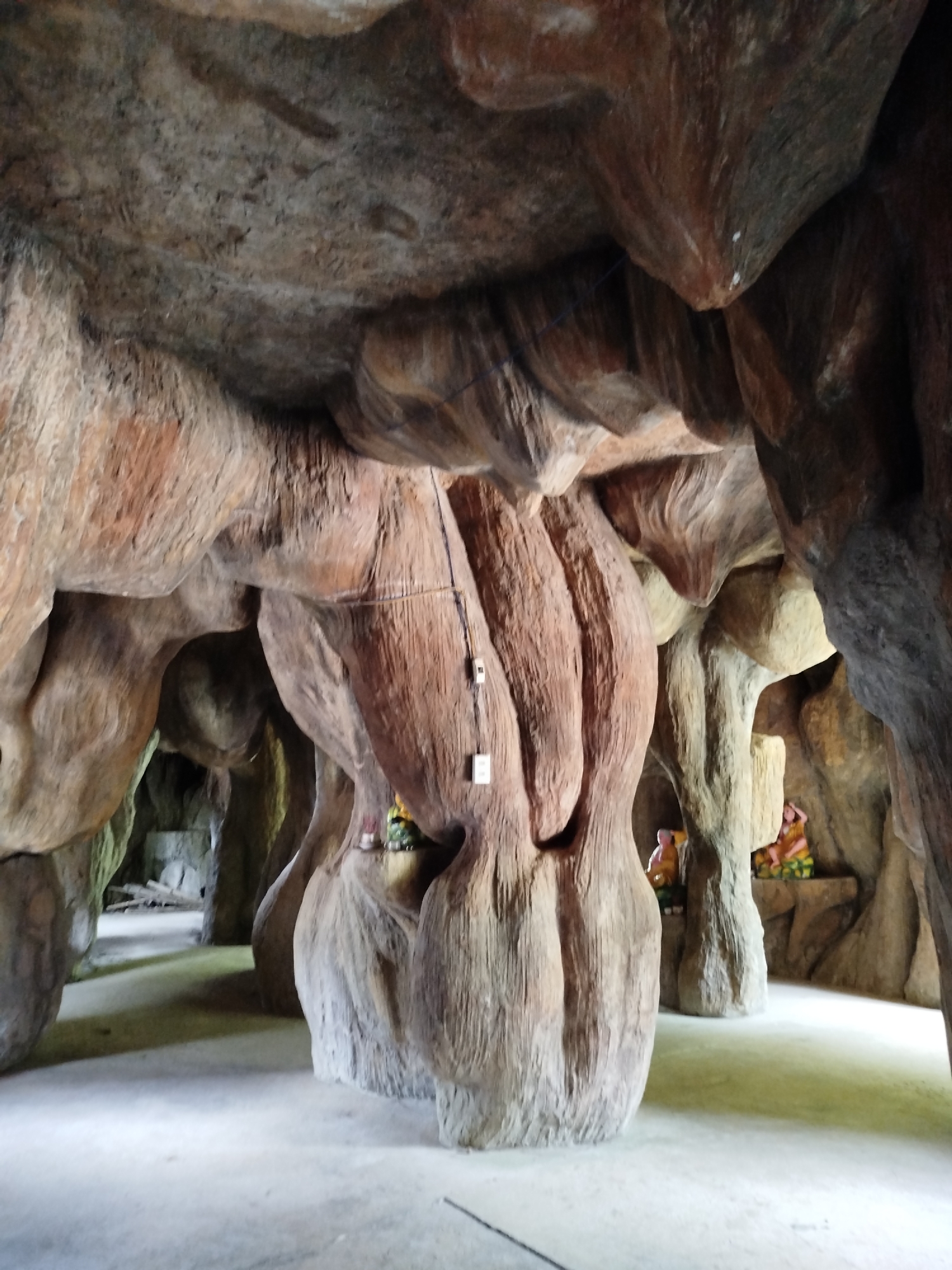 This is a full, eye-level shot of an artificial cave interior with a concrete floor. The cave walls are sculpted to resemble large, gnarled tree trunks and rock formations, with a color palette of browns, grays, and some reddish-brown tones. The ceiling is low and appears to be formed from a single, large, undulating rock-like structure. In the middle ground, a thick, ribbed pillar dominates the center of the frame, with two electrical boxes mounted on its surface. To the right of this pillar, further back in the cave, two small, seated figurines are visible on ledges. The left side of the image shows a pathway leading into the deeper recesses of the cave, with more sculpted rock formations and a hint of green foliage. The lighting is diffused, creating soft shadows. The overall impression is one of a man-made, stylized natural environment.