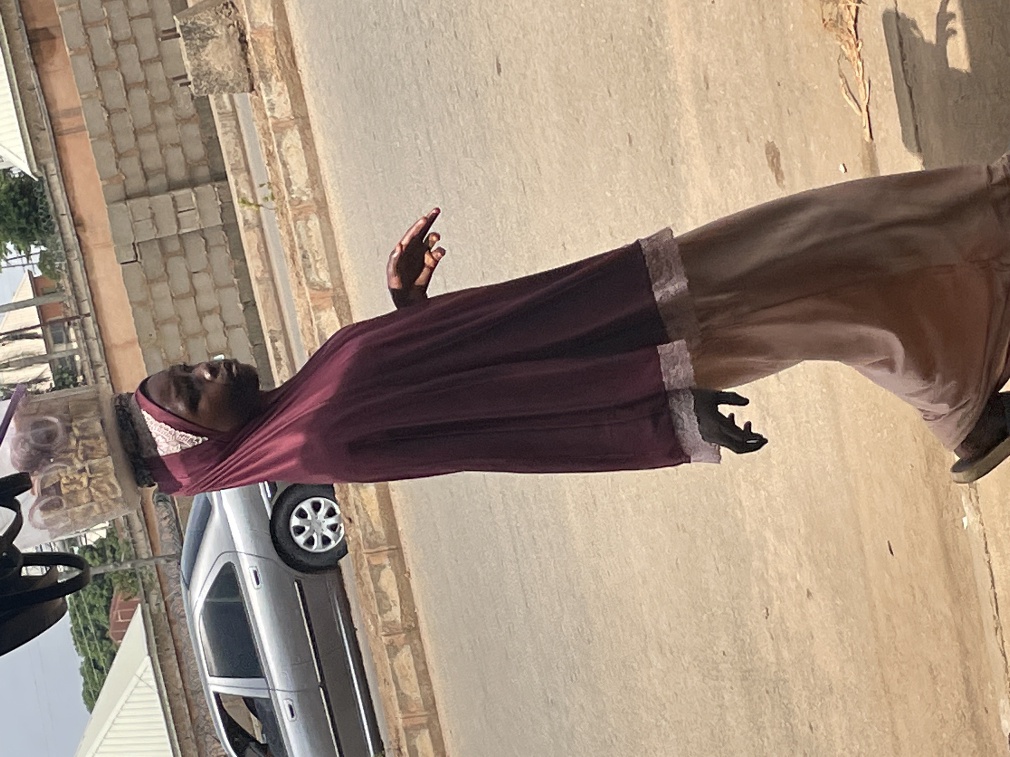 A young Nigerian girl walks along a dirt road in Bukuru, Nigeria, balancing a plastic container filled with food items on her head. She wears a dark red hijab and a light brown skirt, with dark gloves on her hands. A silver sedan is parked on the side of the road in the background, and a brick wall runs along the street. The overall scene depicts a typical day in a Nigerian city.