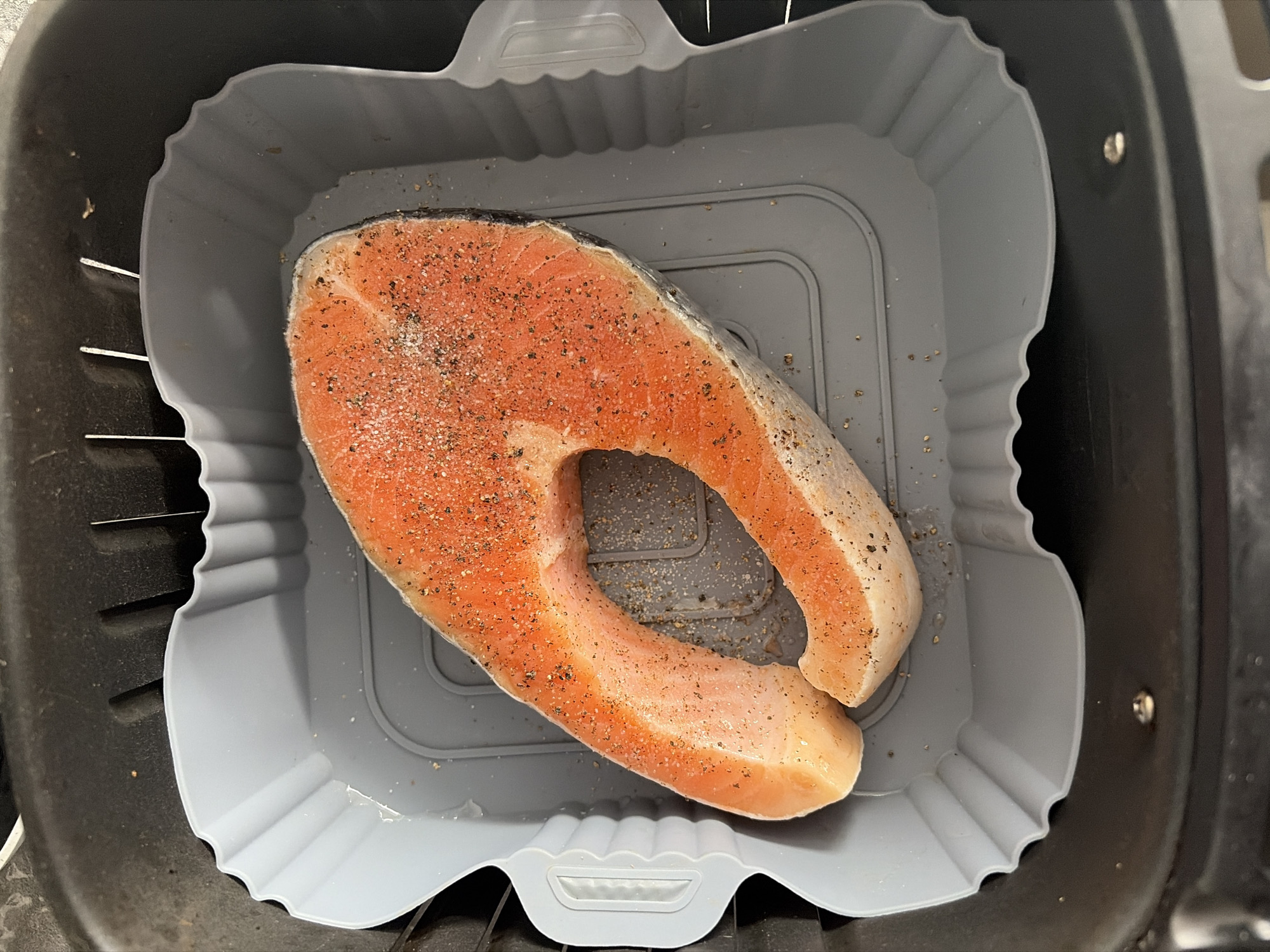 This is a top-down, close-up shot of a single salmon steak, seasoned with salt and pepper, placed inside a grey silicone air fryer liner. The liner has a scalloped edge and sits within a black air fryer basket. The salmon is bright pink and orange with a visible layer of fat. The image is well-lit, suggesting an indoor setting, likely a kitchen. There is no indication of the time of day, weather, or specific location beyond it being in Sochi, Russia, though no distinct visual cues confirm this. The focus is entirely on the food preparation, with no people visible.