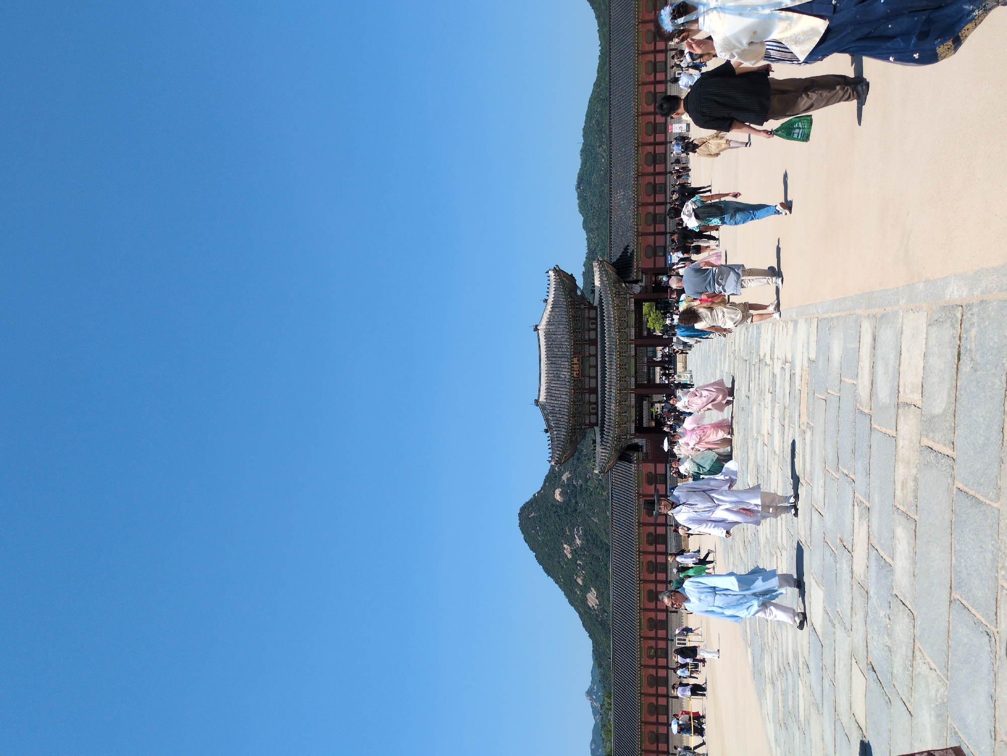 The image shows a crowded walkway at Gyeongbokgung Palace in Seoul, South Korea. The walkway is lined with traditional Korean buildings and a forested hill in the background. Many people dressed in traditional Korean clothing (hanbok) are walking along the walkway. The sky is a clear blue. The ground appears to be paved with stone blocks.
