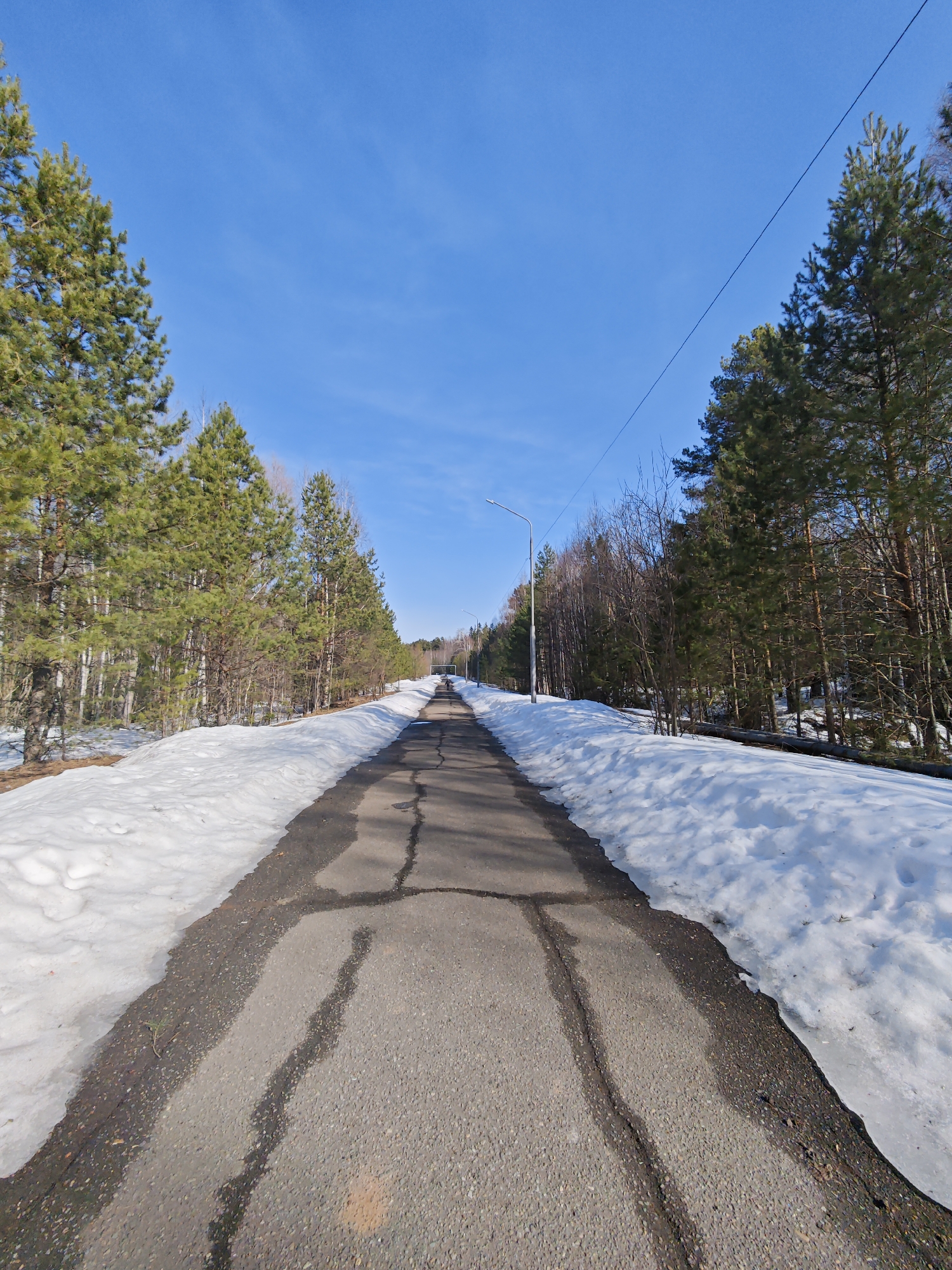 The image depicts a straight, narrow asphalt road stretching into the distance, flanked by snow-covered ground and trees. The road is cracked and shows signs of wear. Trees line both sides of the road, with varying densities of foliage. A clear blue sky is visible above. A utility pole is visible on the right side of the road. The location is Марковский, Russia.